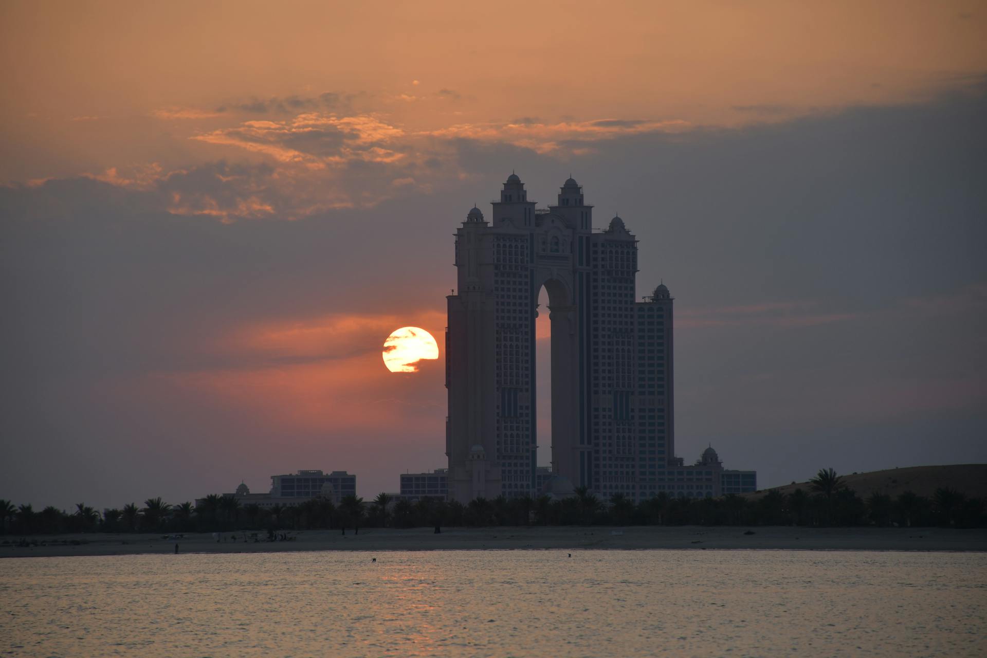 Dramatic sunset behind the Abu Dhabi skyline with a prominent tower silhouette over the water.