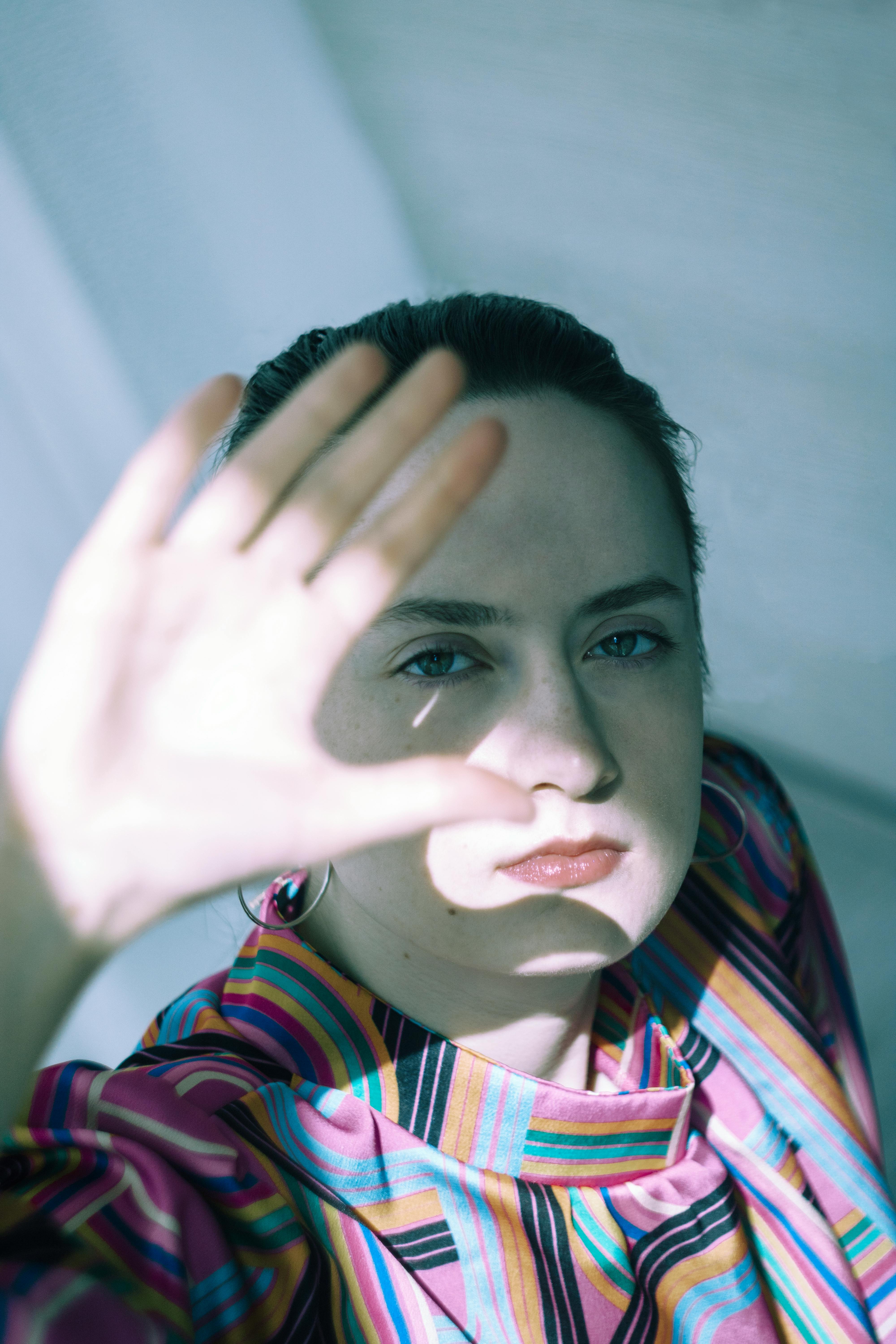 Ücretsiz Güneş ışığını engellemek için elini kaldırmış, canlı çizgili kıyafetler giymiş genç bir kadının portresi. Stok Fotoğraflar