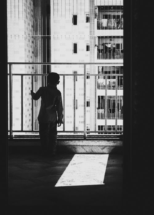 Free A child silhouetted in a dark room gazes out at urban city buildings through a window. Stock Photo