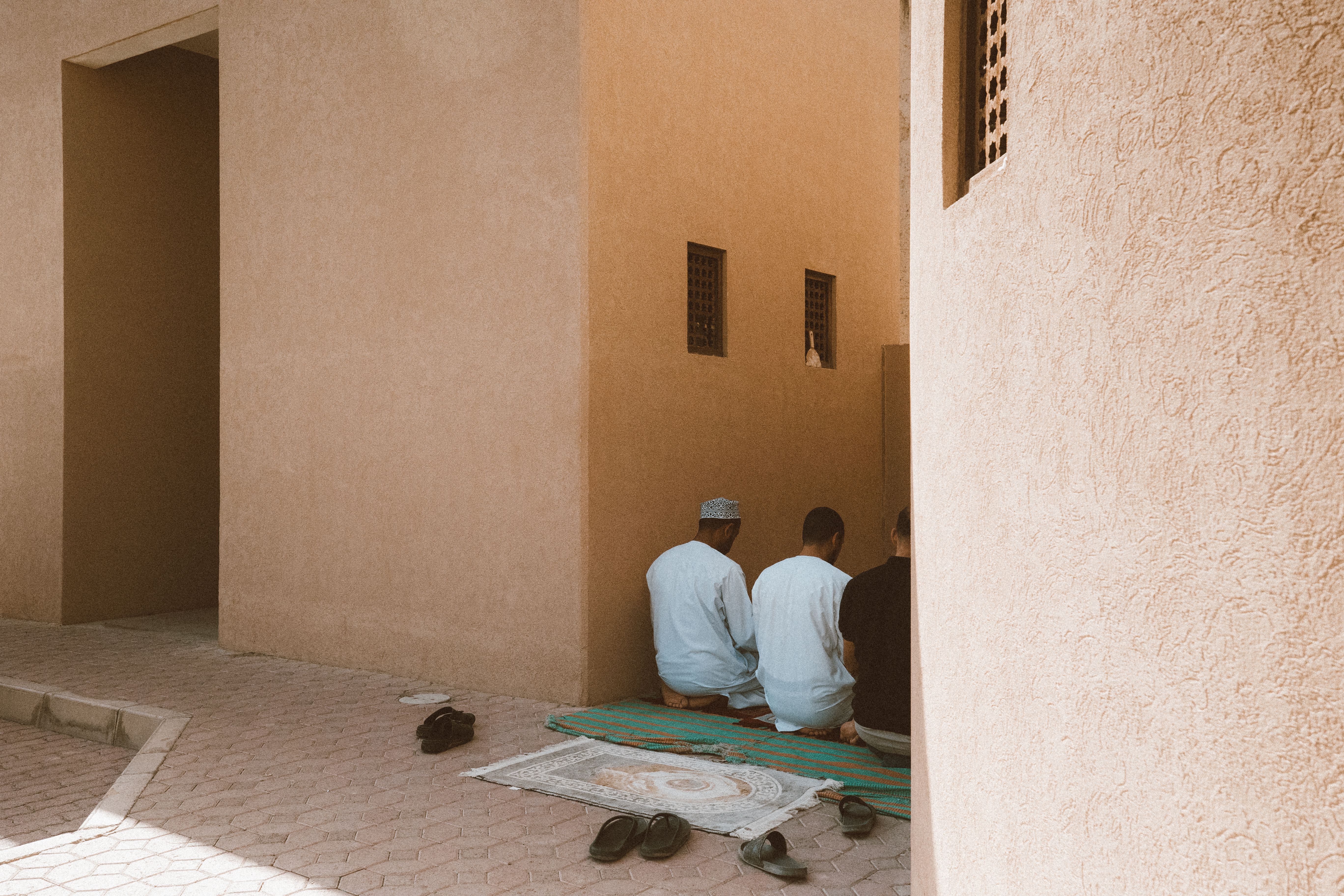 Three men kneeling in prayer outside a traditional Middle Eastern building, surrounded by sand-colored walls.