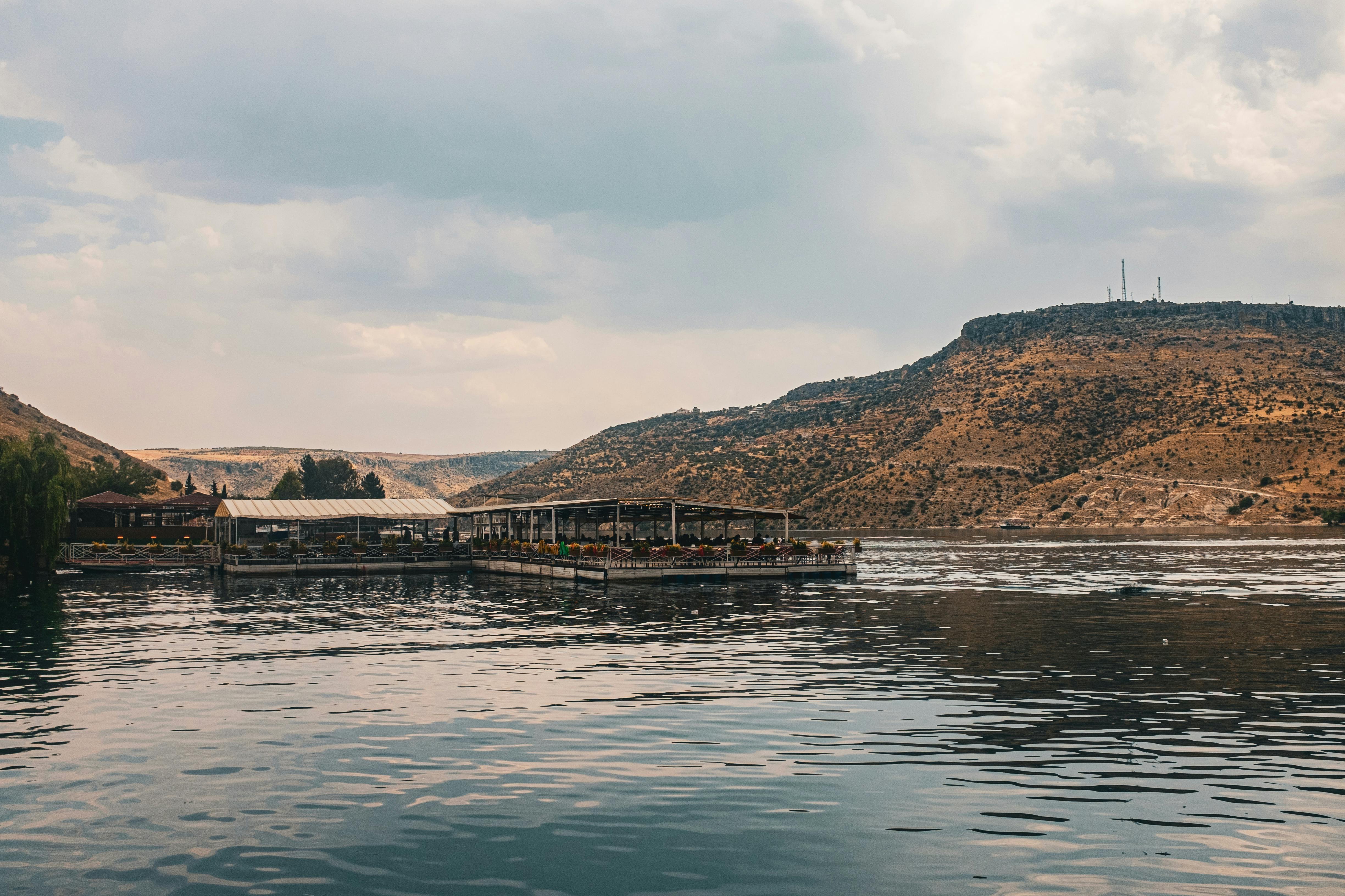 Kostenlos Ein idyllischer Blick auf den See mit einem schwimmenden Restaurant vor der Kulisse von Wüstenhügeln und bewölktem Himmel. Stock-Foto
