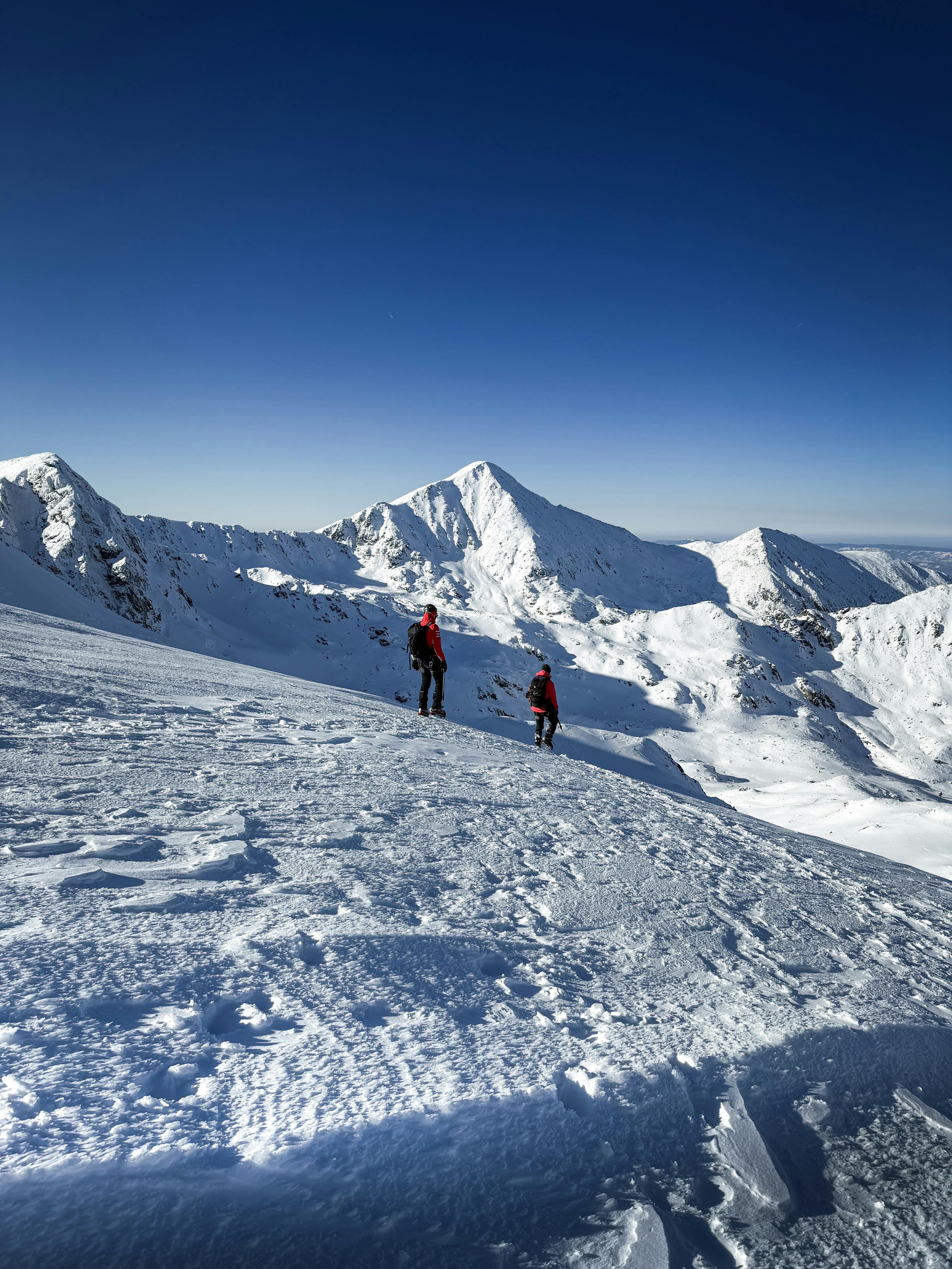 Δωρεάν Δύο πεζοπόροι εξερευνούν ένα χιονισμένο ορεινό τοπίο κάτω από έναν καταγάλανο ουρανό. Φωτογραφία από στοκ φωτογραφιών