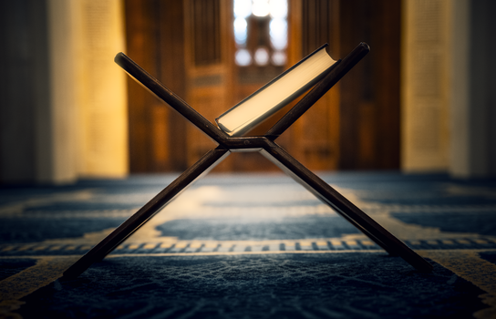 Intricately designed Quran stand inside a mosque with patterned carpet and soft lighting.