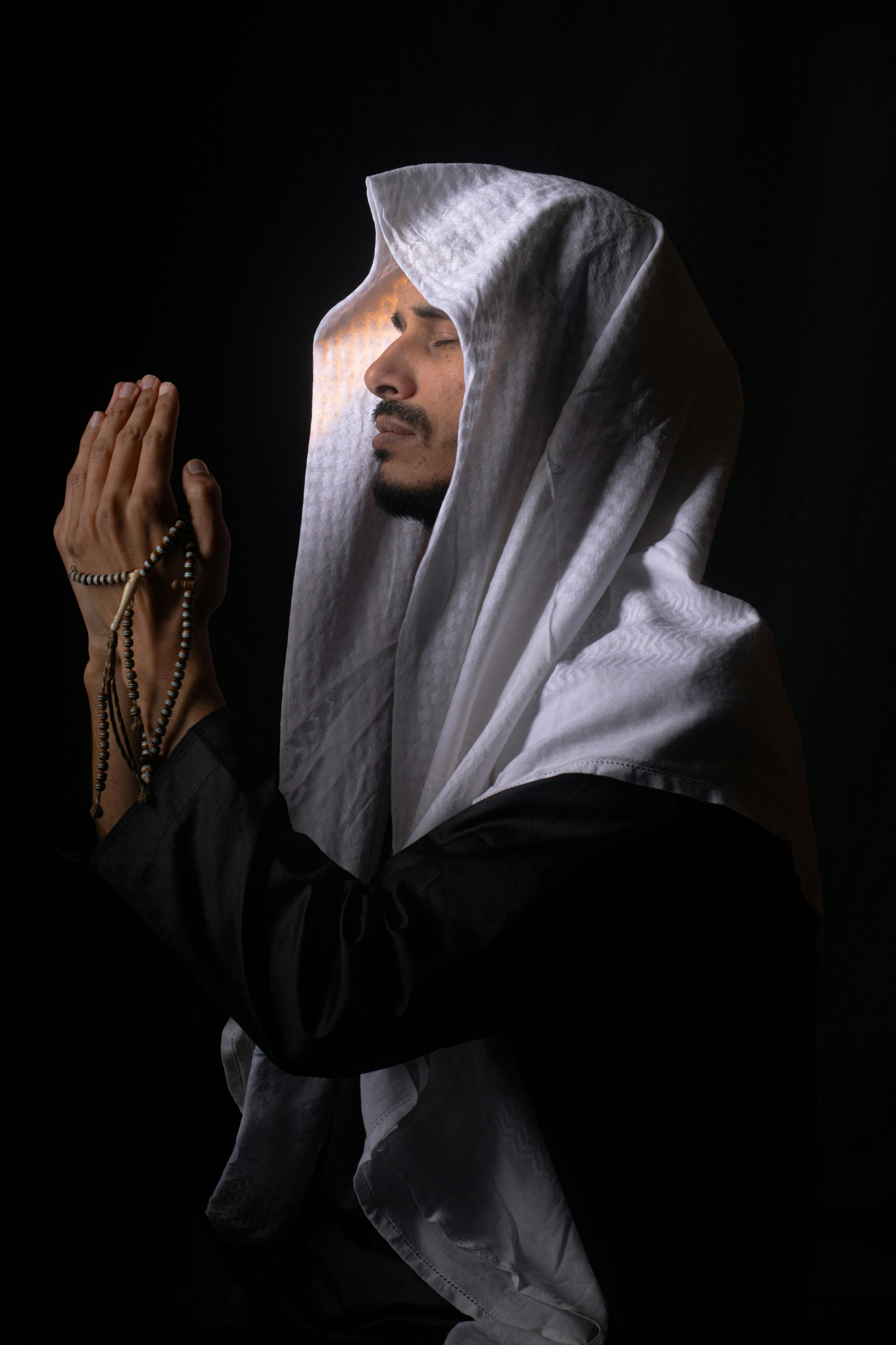 Gratis Ung mann i tradisjonell Midtøsten-antrekk ber, med rosenkransperler mot en mørk bakgrunn. Arkivbilde