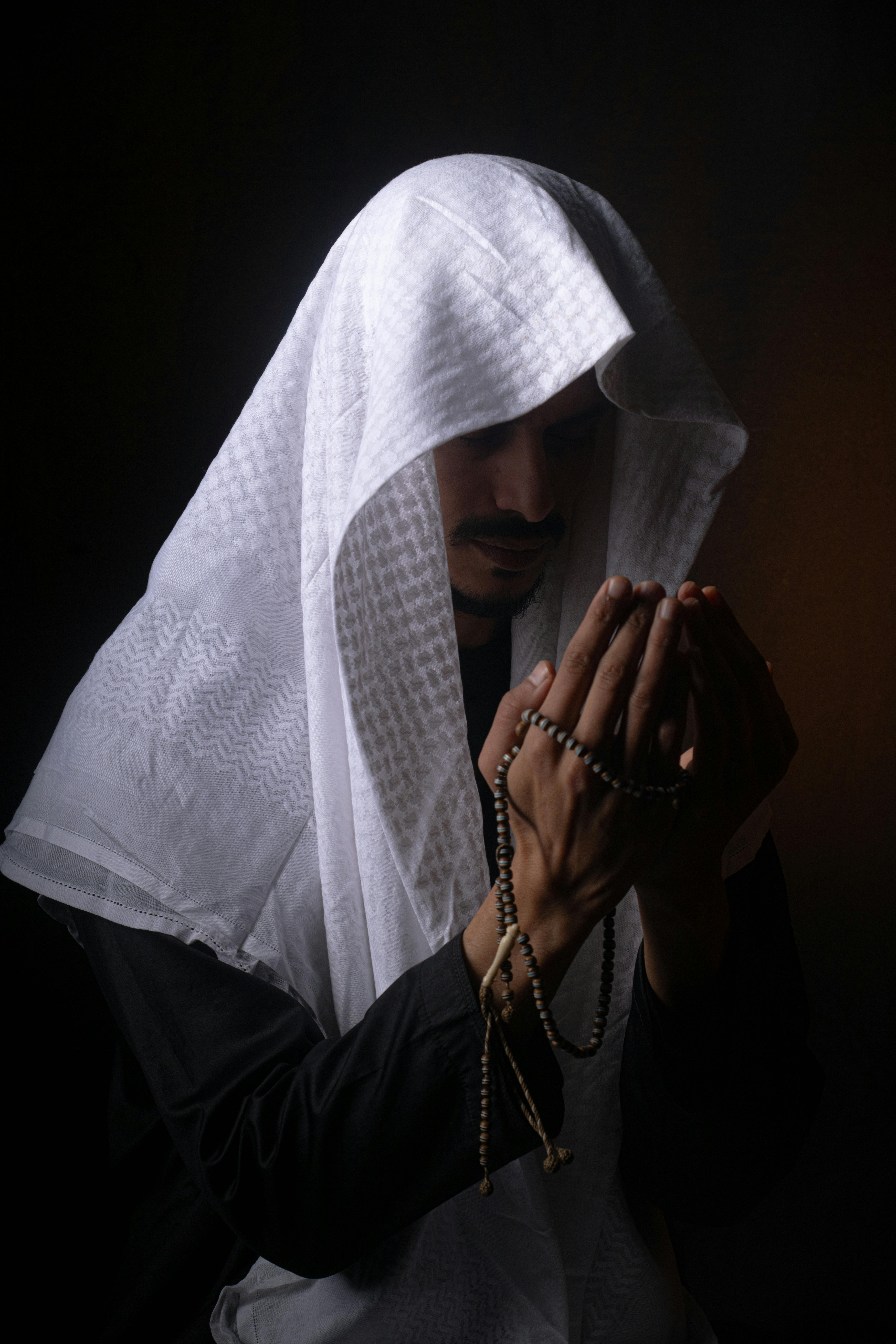 Kostenlos Ein Mann aus dem Nahen Osten in traditioneller Kleidung betet in dunkler Umgebung mit einem Rosenkranz. Stock-Foto