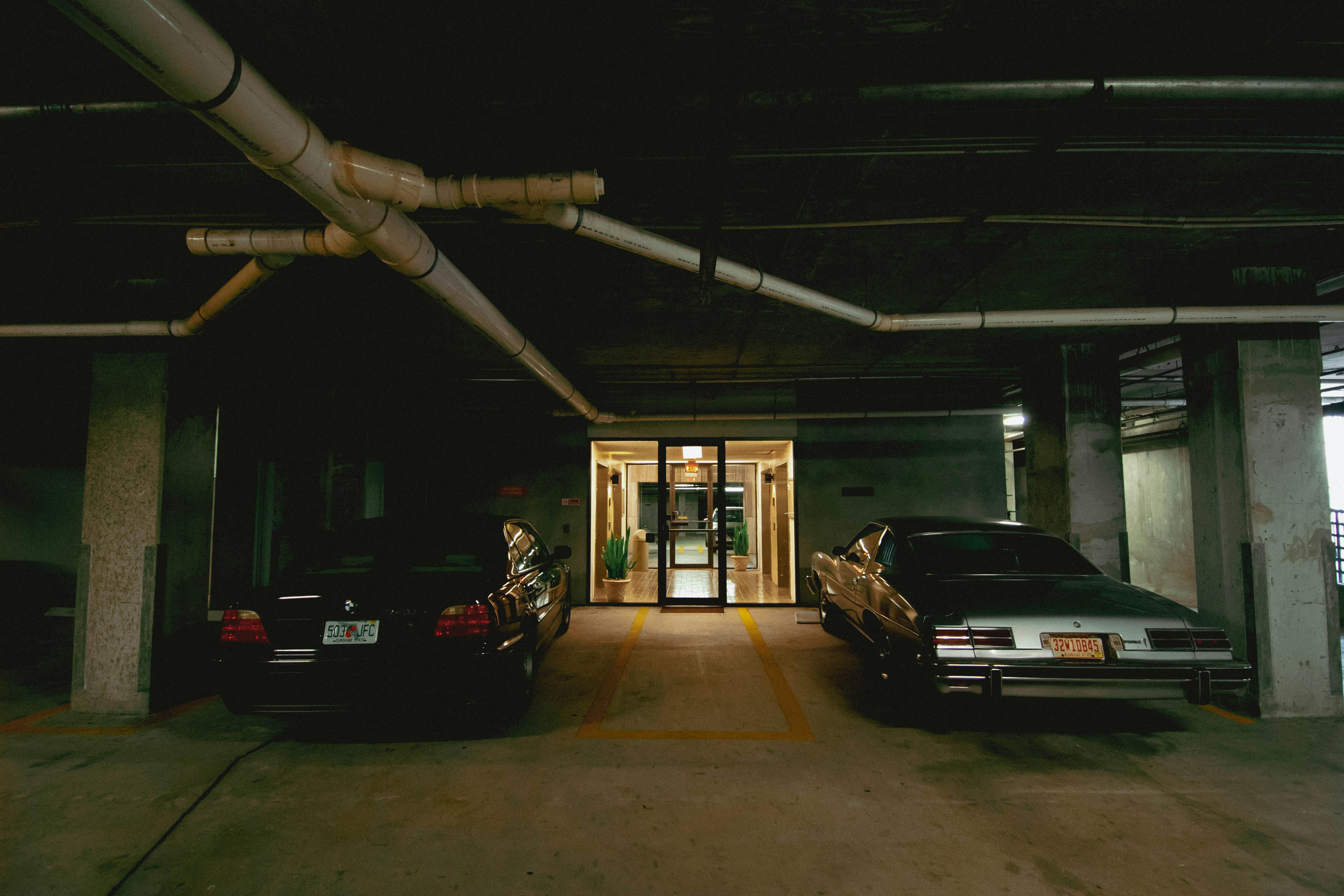 Gratis To biler parkert i et svakt opplyst, underjordisk parkeringshus med synlige rør. Arkivbilde