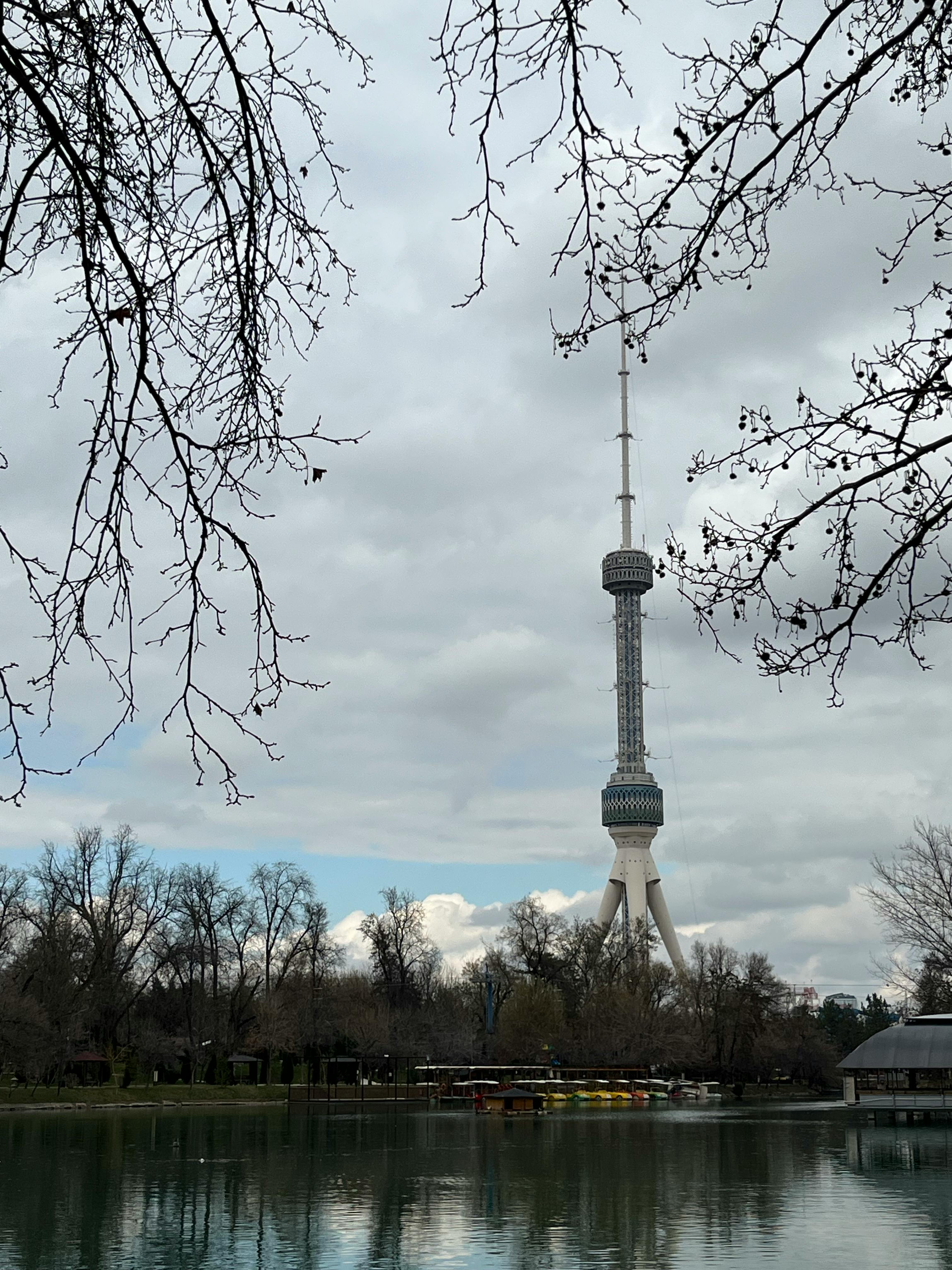 Kostenlos Der Taschkenter Turm vor bewölktem Himmel, umgeben von Bäumen und Spiegelungen in einer idyllischen Parklandschaft. Stock-Foto