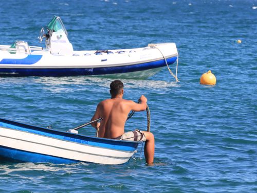 Free A young man rows a small boat in a clear blue sea, with a larger boat in the background. Stock Photo