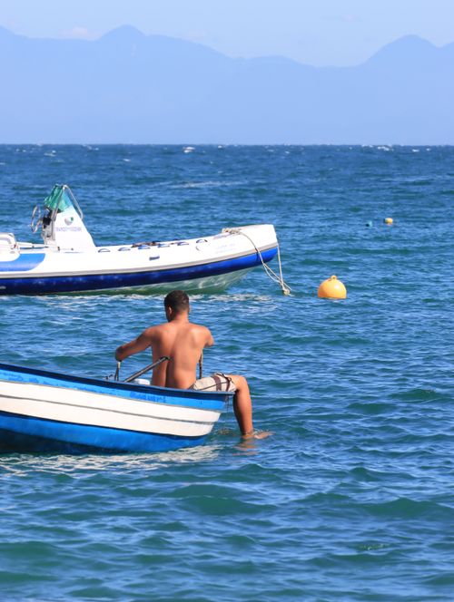 Free A man enjoys a peaceful day on the ocean in a small boat with a distant mountain view, under a clear blue sky. Stock Photo