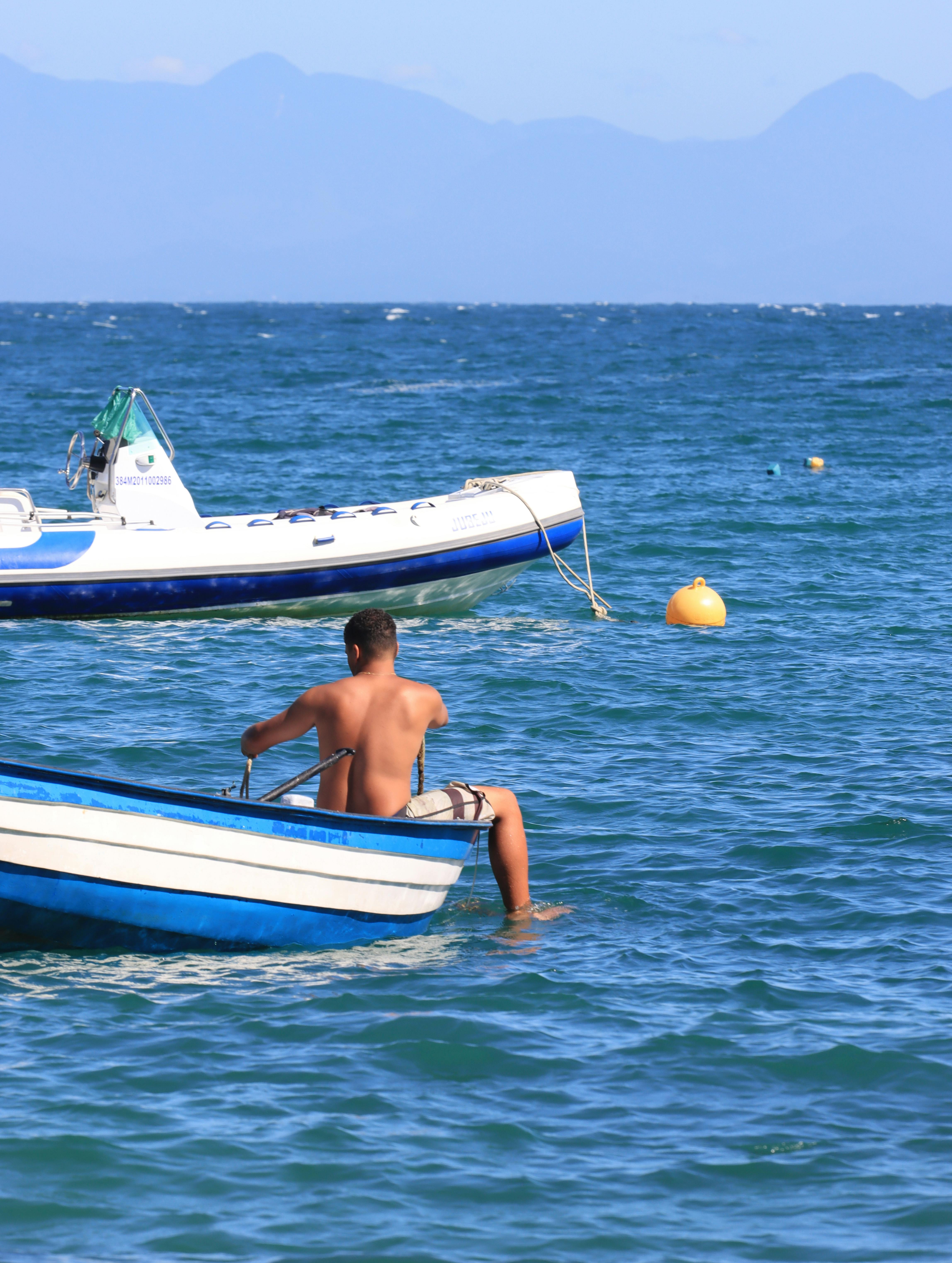 Kostenlos Ein Mann genießt einen friedlichen Tag auf dem Meer in einem kleinen Boot mit Blick auf die fernen Berge unter einem klaren blauen Himmel. Stock-Foto