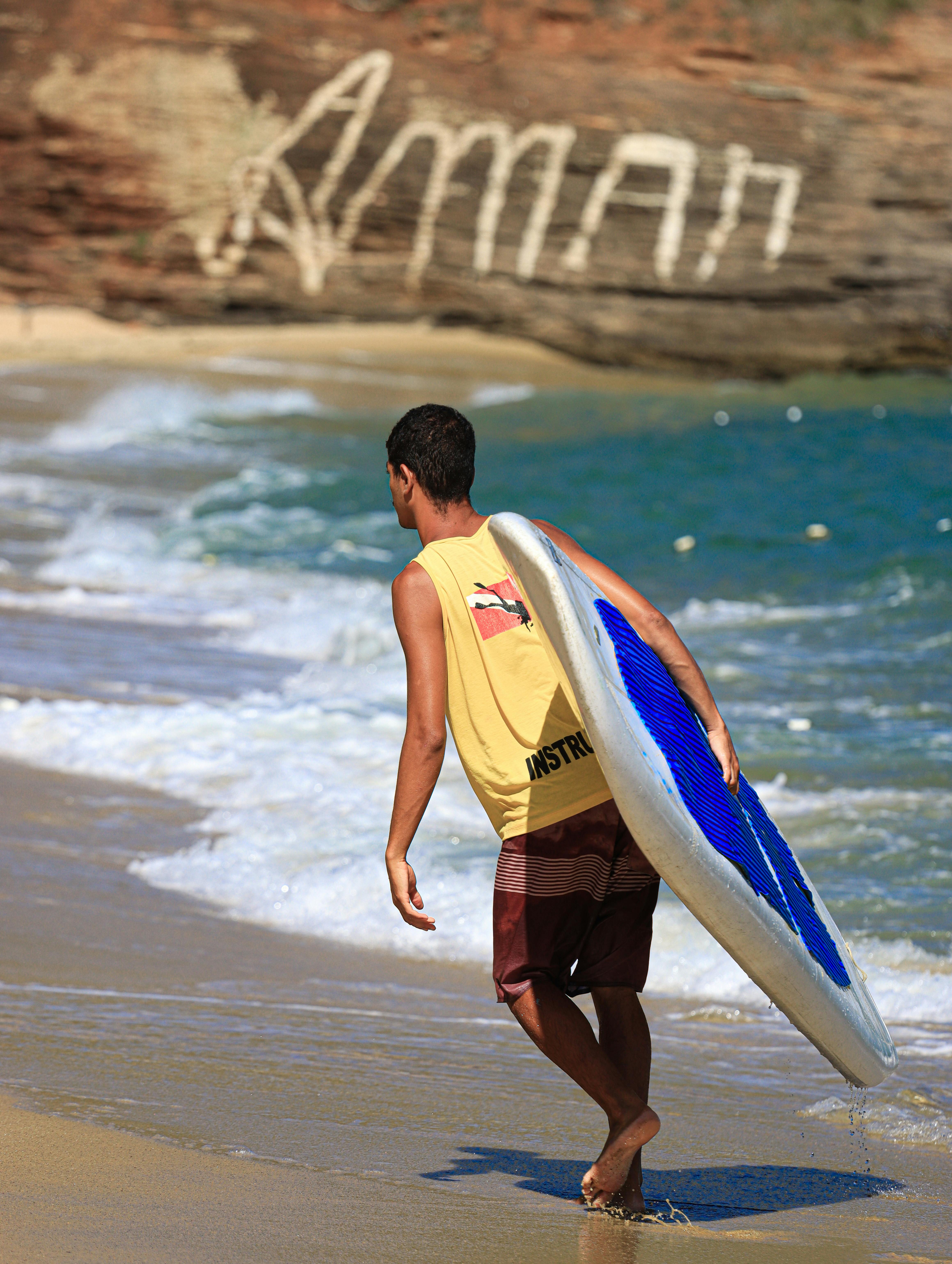 Kostenlos Ein Mann spaziert mit einem Surfbrett unter strahlend blauem Himmel an einem Sandstrand entlang. Eine perfekte Szene für ein Sommerabenteuer. Stock-Foto
