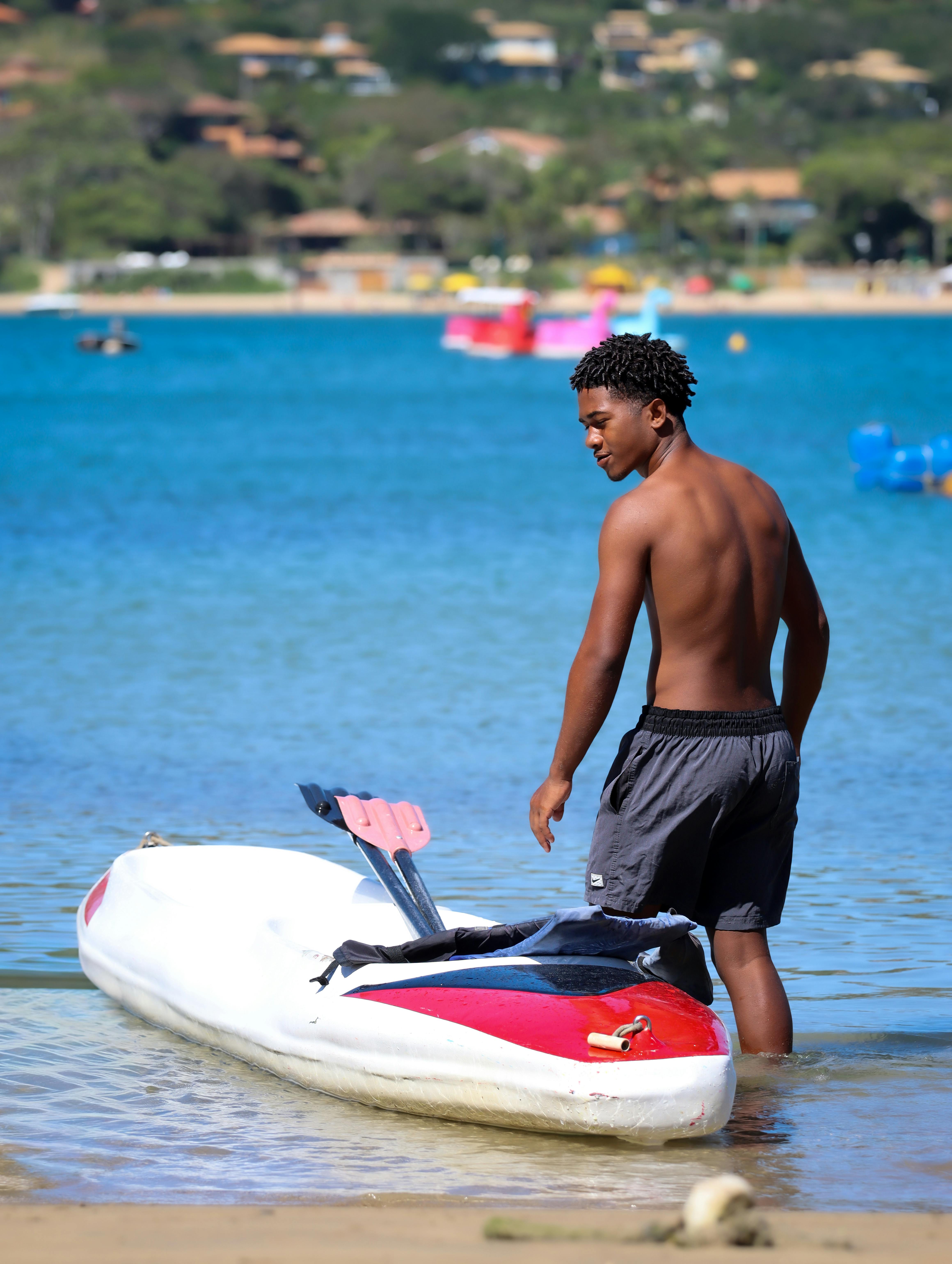 Gratis En ung mand står ved en kajak på stranden og nyder en solskinsdag nær vandkanten. Lagerfoto