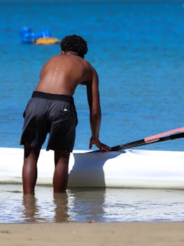 A man adjusts his kayak by the beach, under a bright daytime sky, ready to paddle.