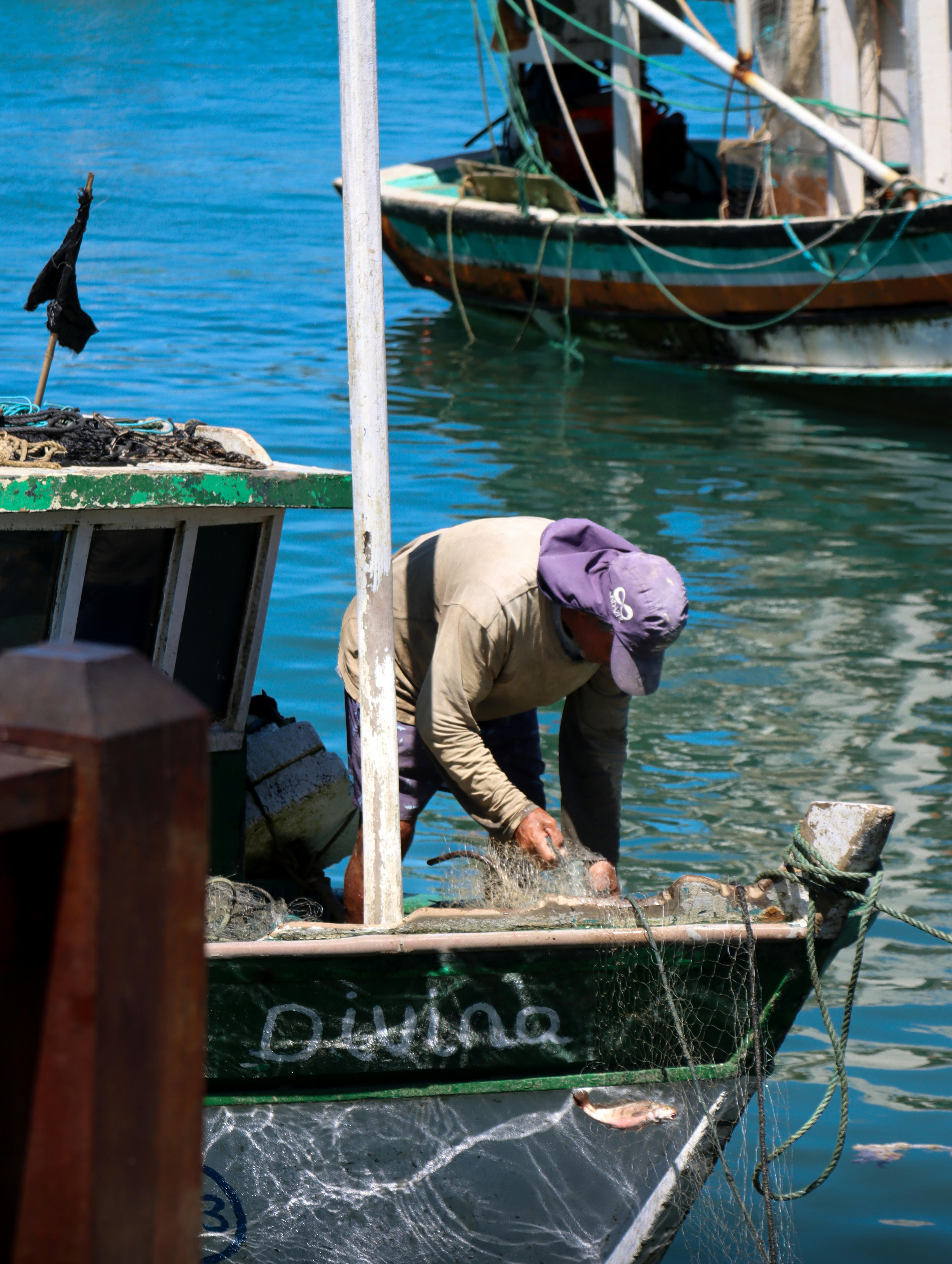 Gratis En fisker reparerer sit net på en lille fiskerbåd, der ligger fortøjet ved det blå vand. Lagerfoto