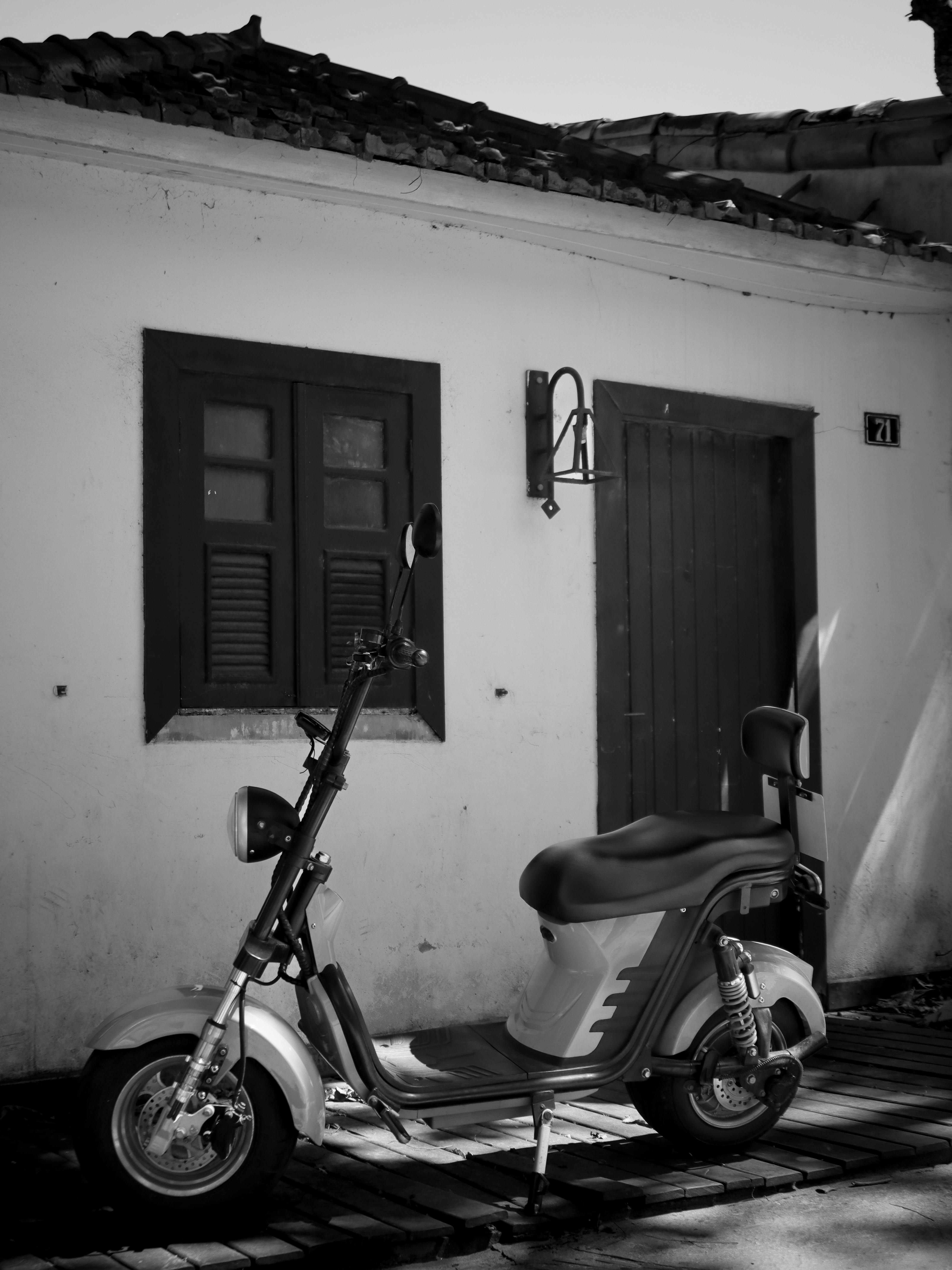 無料 素朴な家の正面を背景にしたクラシックなスクーターの白黒画像。ノスタルジックな雰囲気を醸し出しています。 写真素材