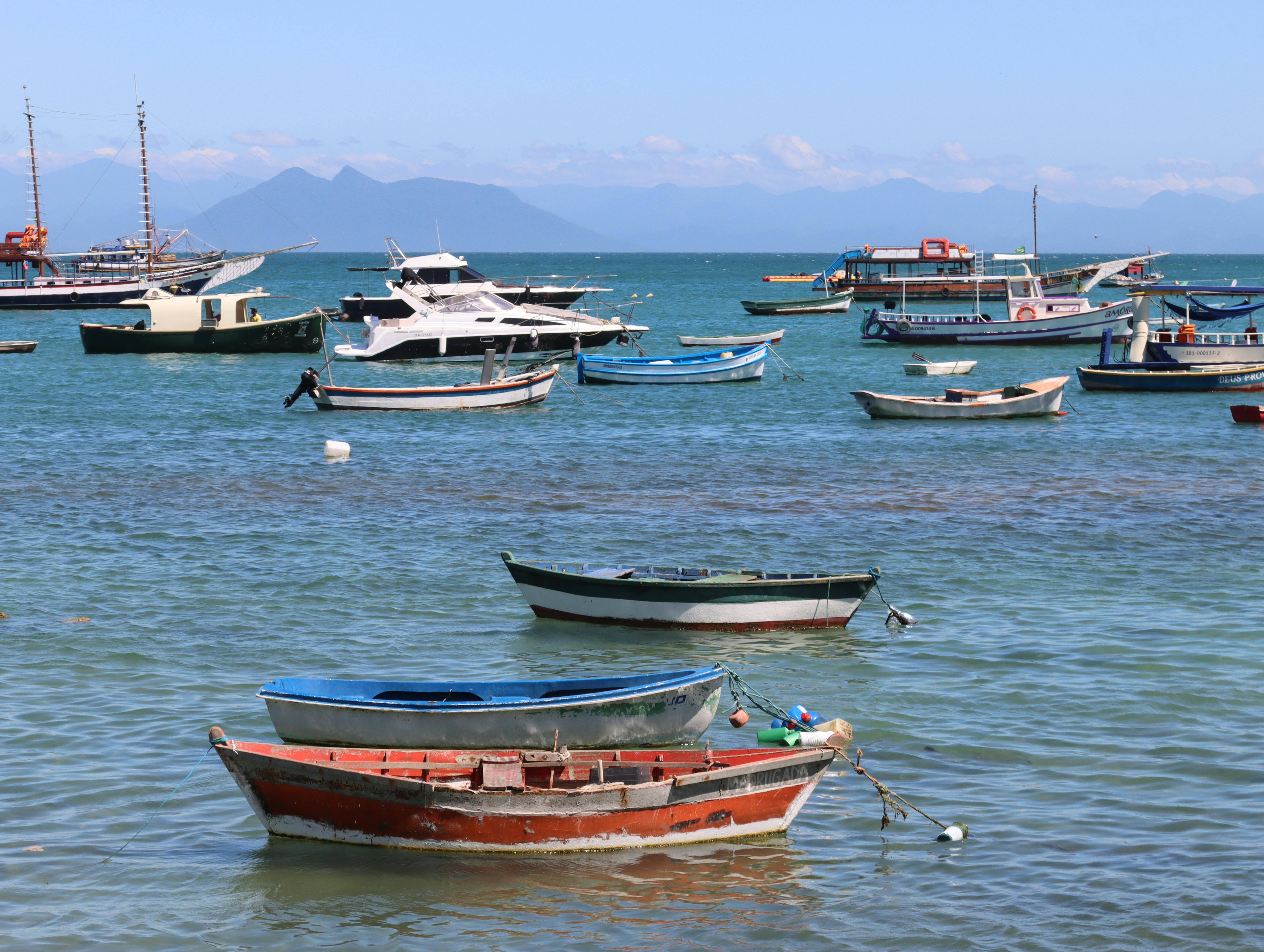 Gratis Farverige fiskerbåde forankret i en fredfyldt bugt med fjerne bjerge under en klar blå himmel. Lagerfoto
