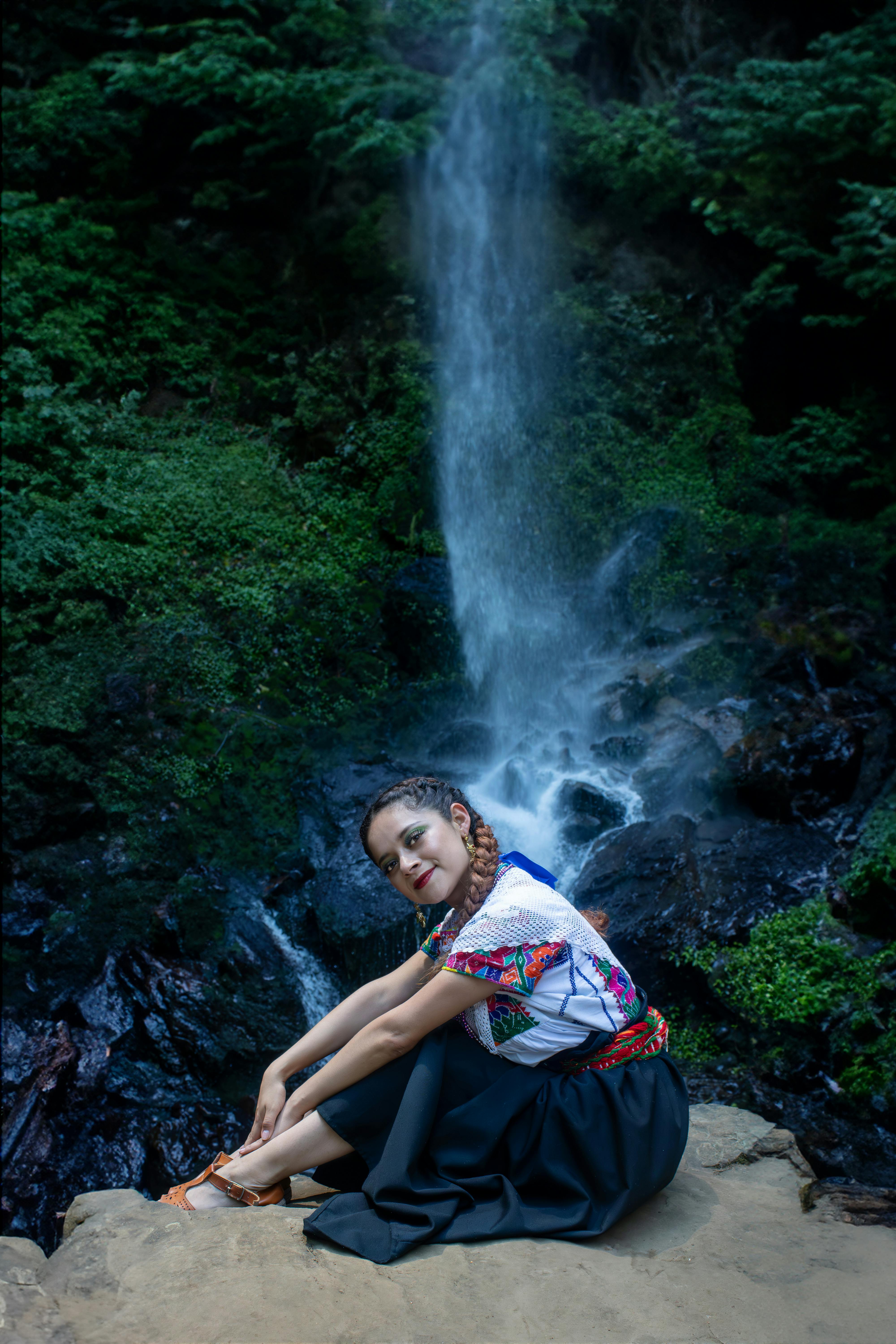 Kostenlos Eine junge Frau sitzt an einem Wasserfall, umgeben von üppigem Grün, in einer idyllischen Naturlandschaft. Stock-Foto
