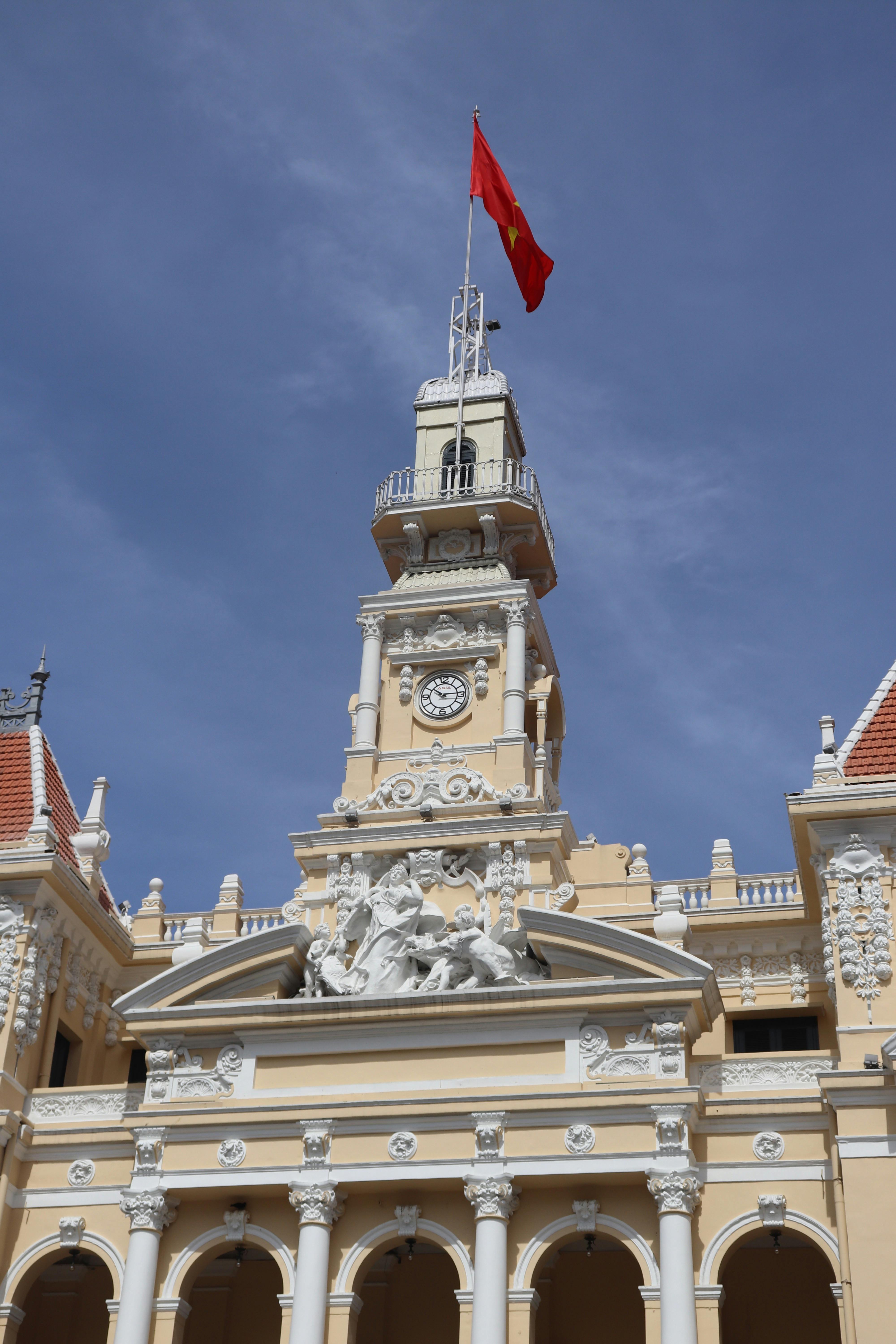 Kostenlos Die elegante Architektur des Rathauses von Ho-Chi-Minh-Stadt mit der vietnamesischen Flagge unter strahlend blauem Himmel. Stock-Foto