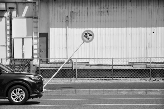 Black and white image of a leaning traffic sign beside a parked car and industrial wall.