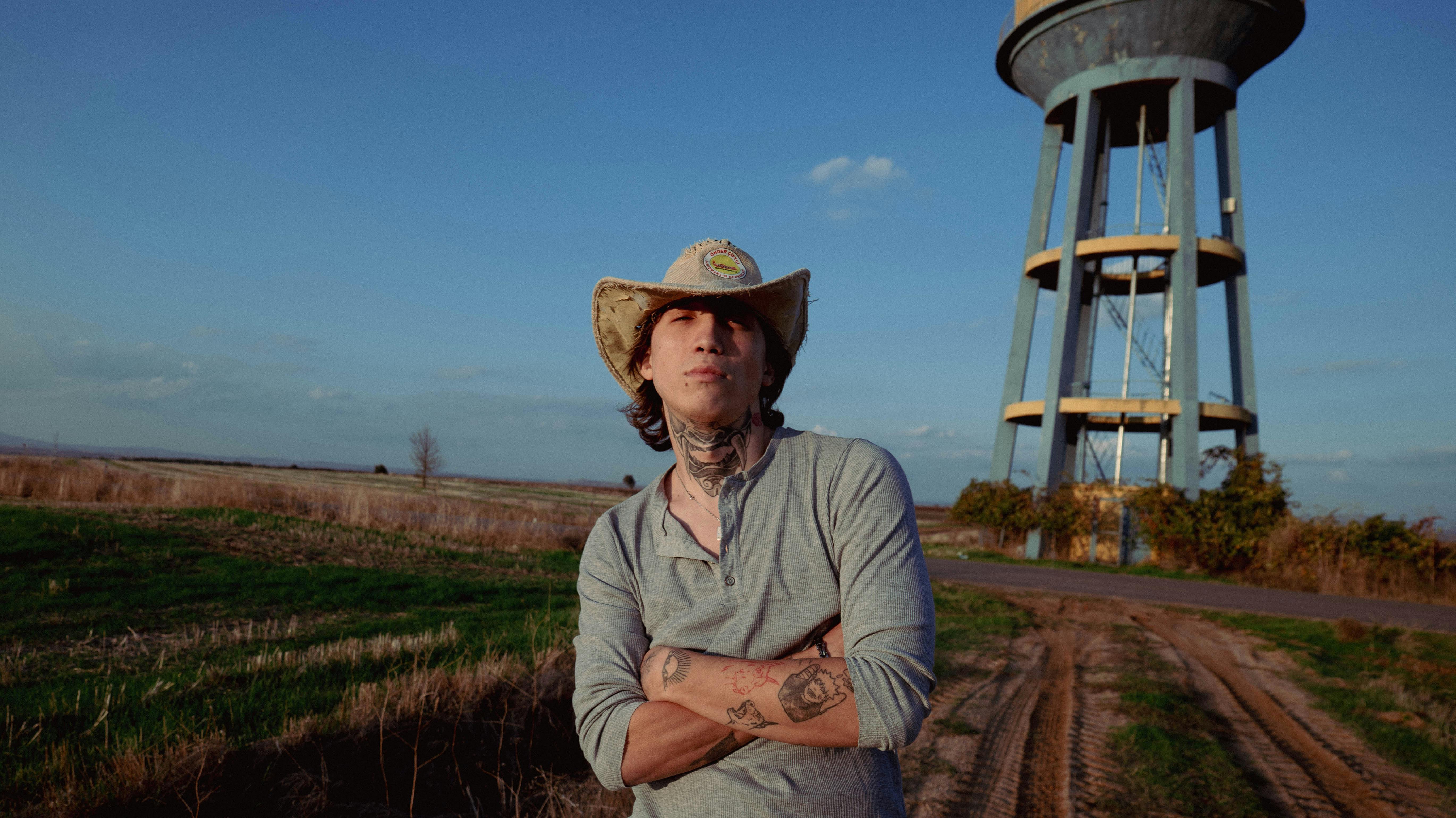 Gratis Portrett av en ung mann i cowboyhatt med tatoveringer, stående ved et vanntårn i et landlig landskap. Arkivbilde