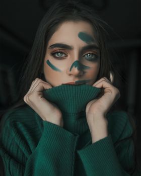 Captivating artistic portrait of a woman with face paint, featuring a green sweater in a studio setting.