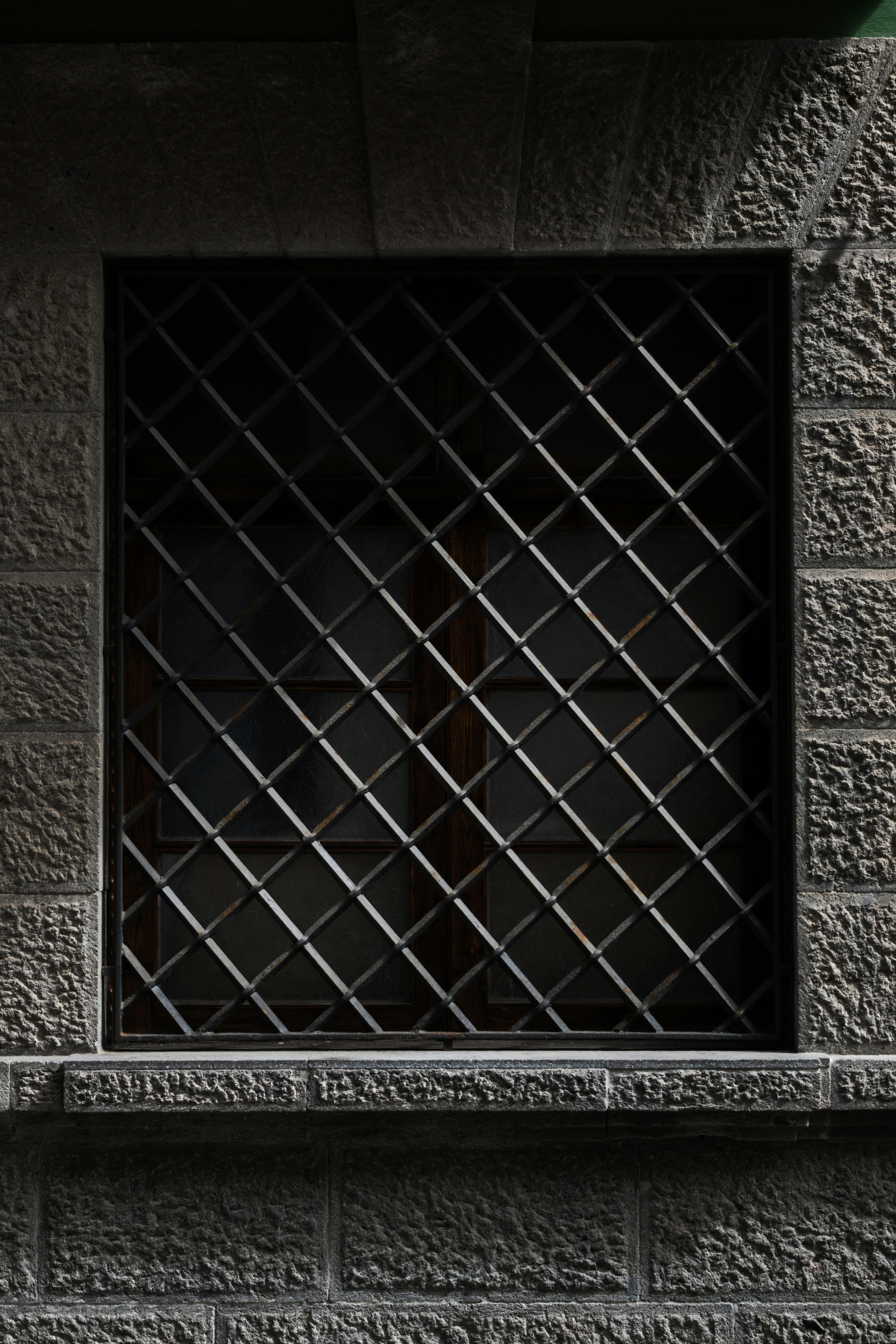 Free A textured stone wall partially shadowed, featuring a window with an intricate iron grate. Stock Photo