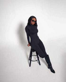 Stylish woman in black sitting on a stool in a minimalist studio setting.