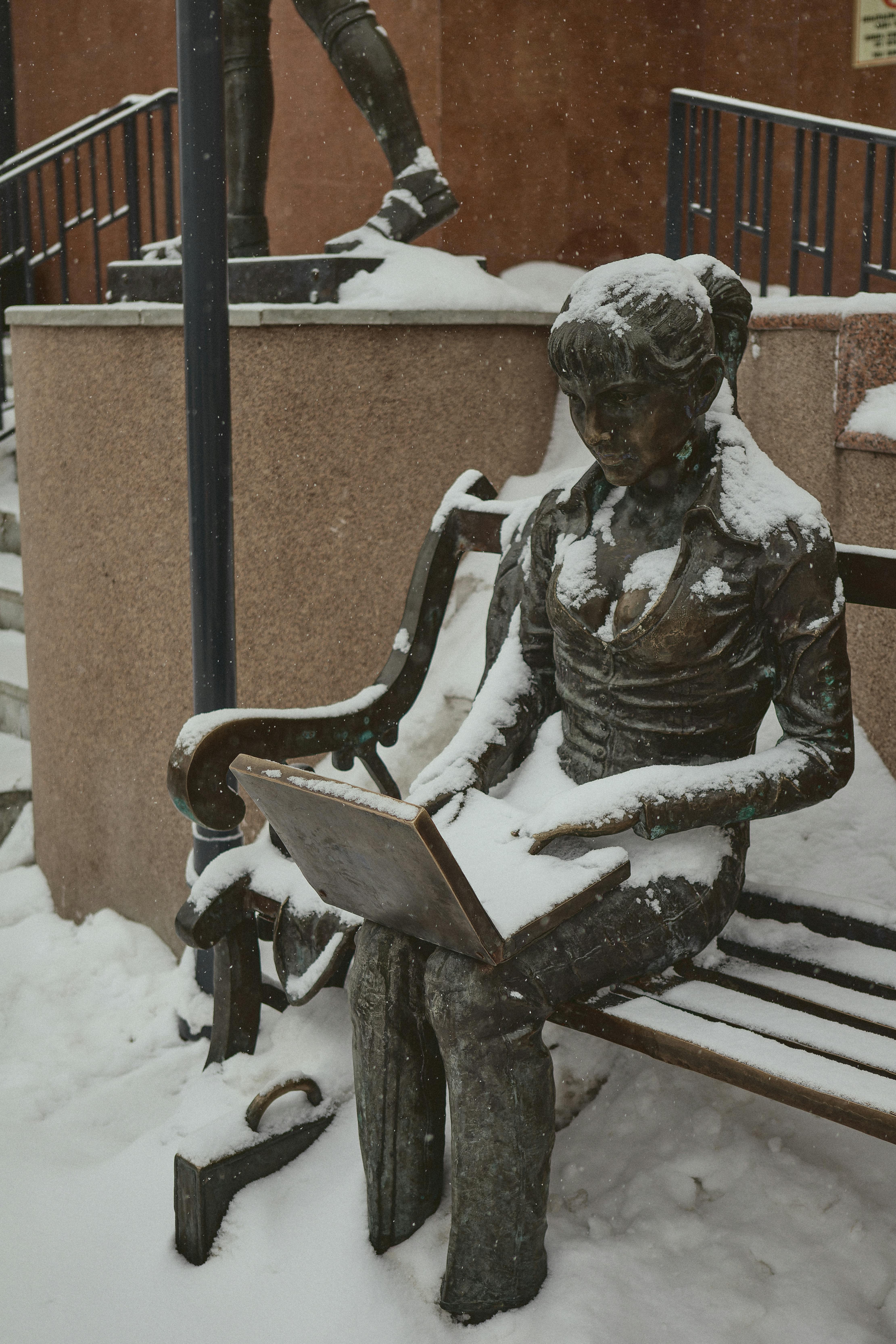 Kostenlos Bronzestatue auf einer schneebedeckten Bank, die einen Laptop hält, erzeugt eine winterliche Stadtszene. Stock-Foto
