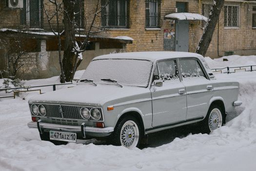 A vintage car covered in snow parked in front of an urban residential building.