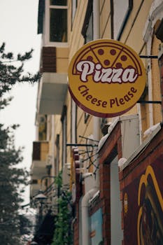 Urban pizza shop sign reading 'Pizza Cheese Please' on a snowy day.