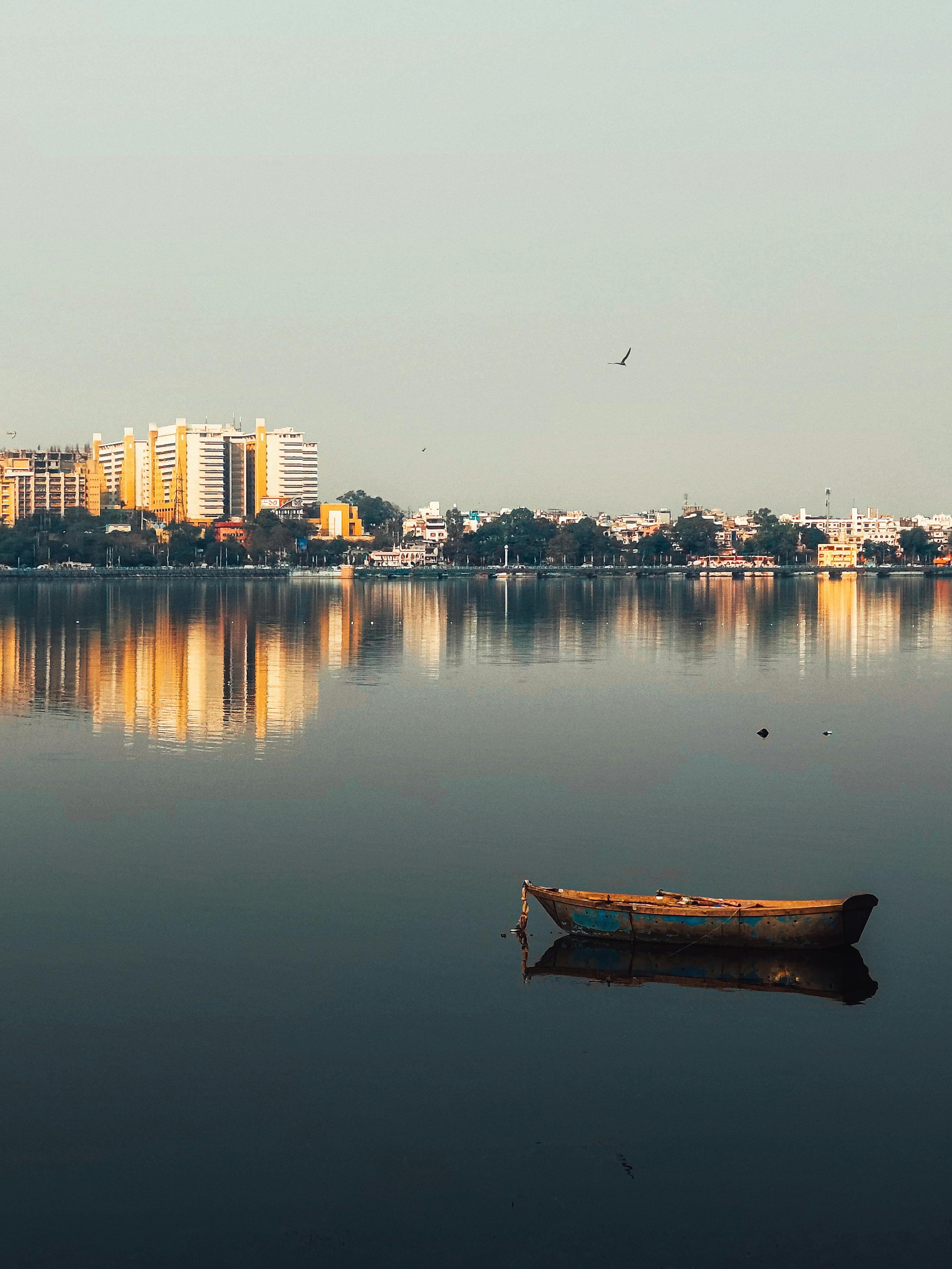 Kostenlos Ein friedlicher Anblick: Ein einsames Boot auf einem ruhigen Stadtsee bei Sonnenaufgang spiegelt die Gebäude wider. Stock-Foto