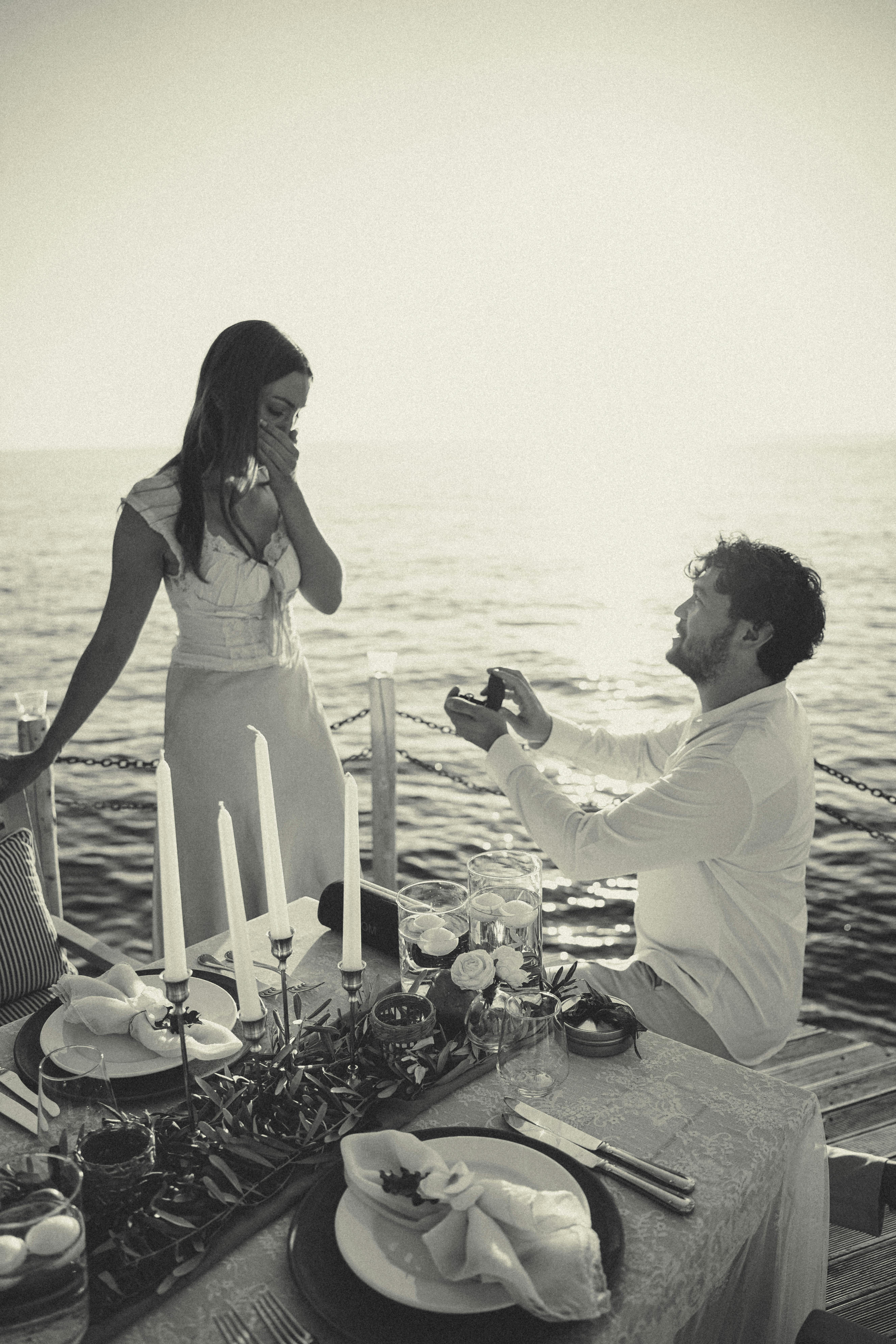 Kostenlos Ein Mann macht einer Frau bei Sonnenuntergang am Meer, umgeben von einer romantisch gedeckten Tafel, einen Heiratsantrag. Stock-Foto