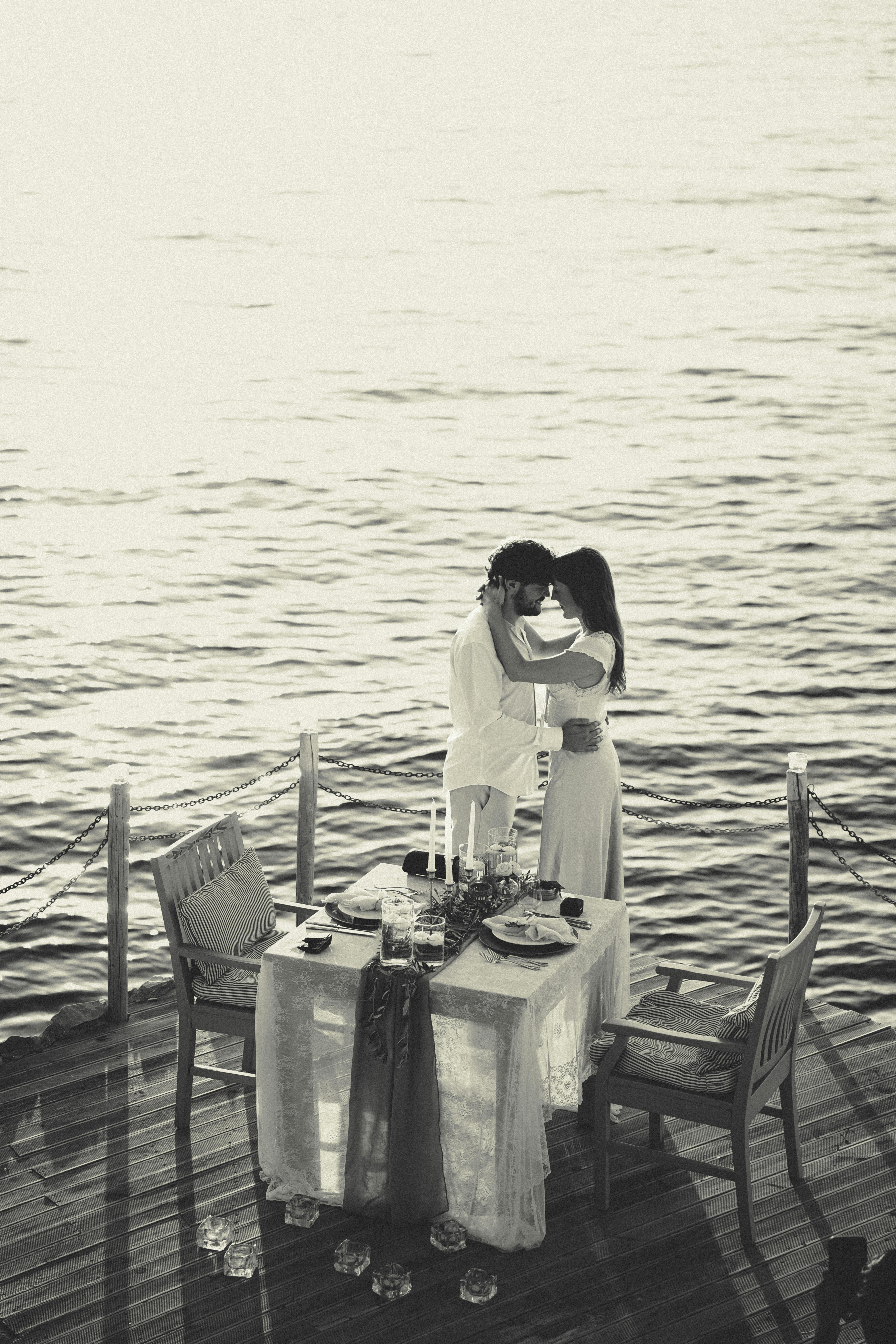 Kostenlos Ein romantisches Schwarz-Weiß-Foto eines Paares, das sich am Meer umarmt, während auf einer Holzterrasse ein Abendessen gedeckt ist. Stock-Foto