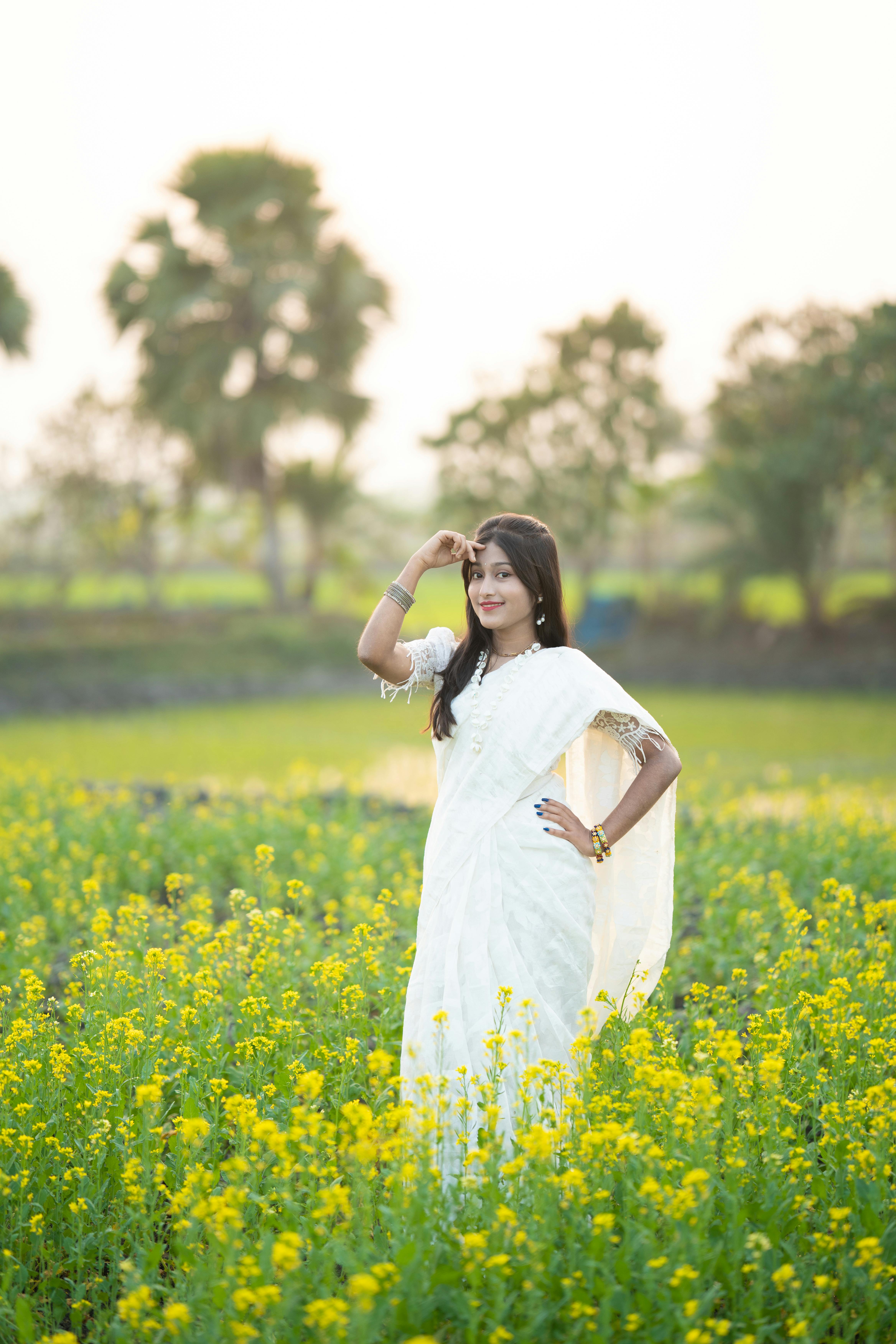 Kostenlos Eine Frau posiert anmutig in einem leuchtend gelben Blumenfeld und trägt einen traditionellen weißen Sari. Stock-Foto