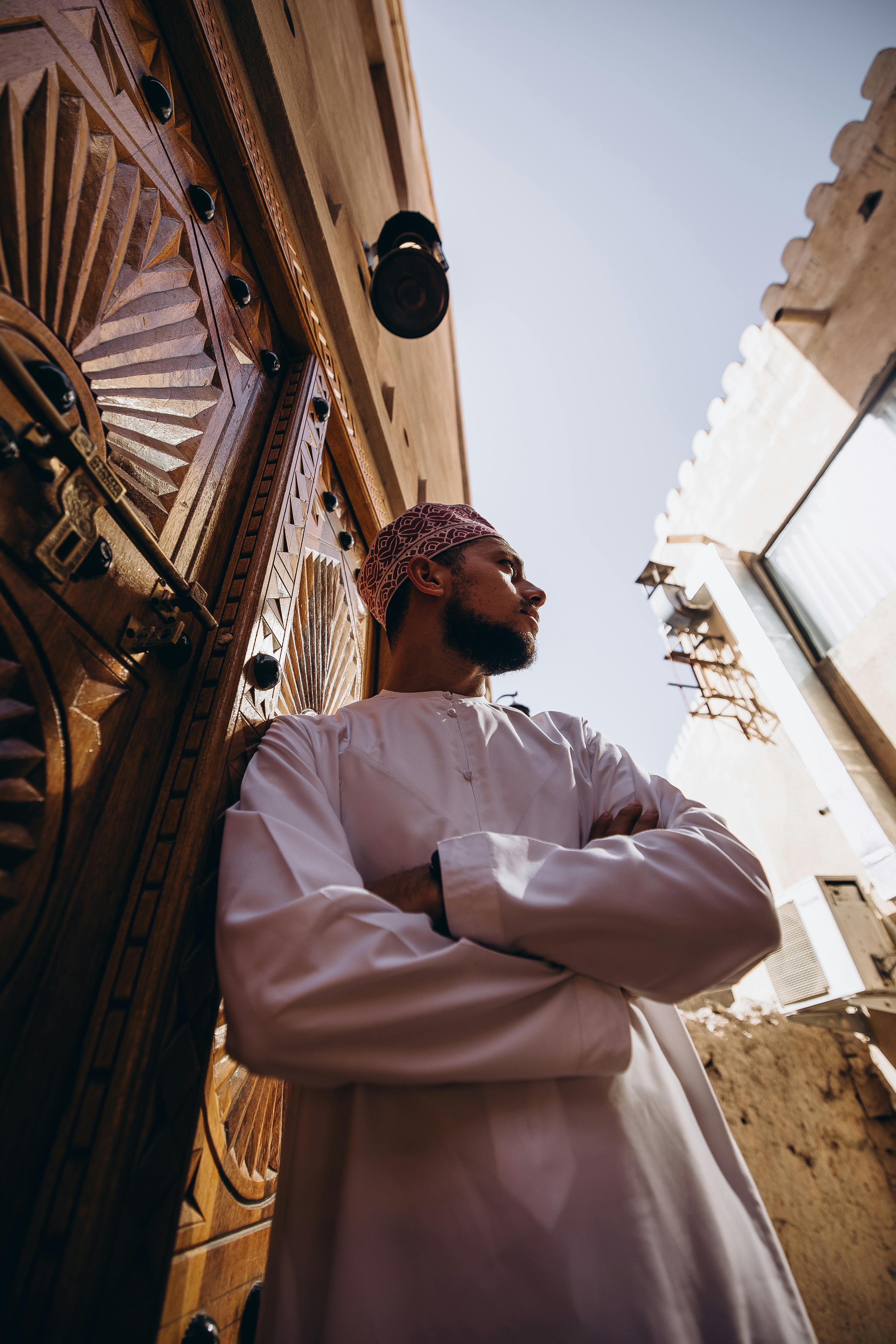 Kostenlos Porträt eines omanischen Mannes in traditioneller Kleidung, der vor einer kunstvoll verzierten Holztür in Nizwa, Oman, steht. Stock-Foto