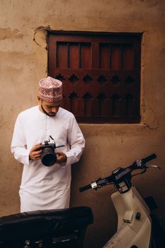 An Omani photographer holds a camera in front of rustic architecture, dressed in traditional attire with a scooter nearby.
