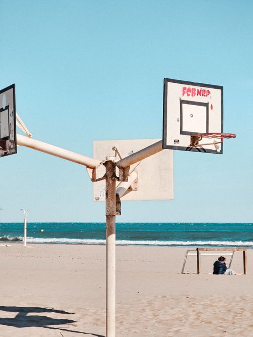 Free A basketball hoop stands on a sandy beach by the ocean under a clear blue sky with a person sitting nearby. Stock Photo