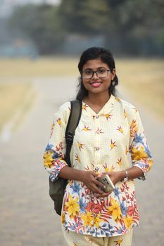 Cheerful young woman with glasses holding phone outdoors in Kolkata, India.
