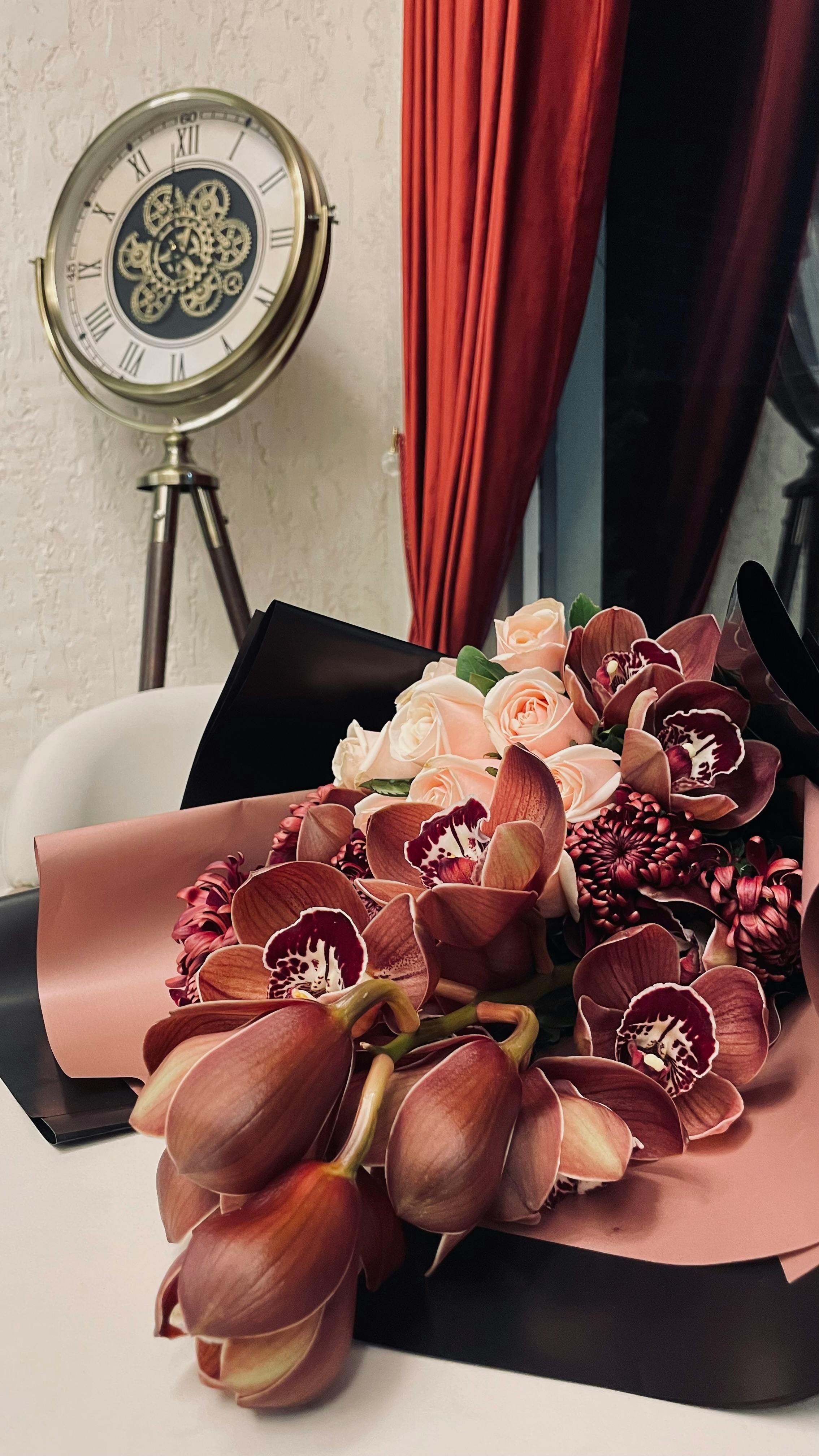 Δωρεάν Πολυτελές μπουκέτο από ορχιδέες και τριαντάφυλλα σε ένα τραπέζι, με vintage ρολόι και κόκκινες κουρτίνες. Φωτογραφία από στοκ φωτογραφιών