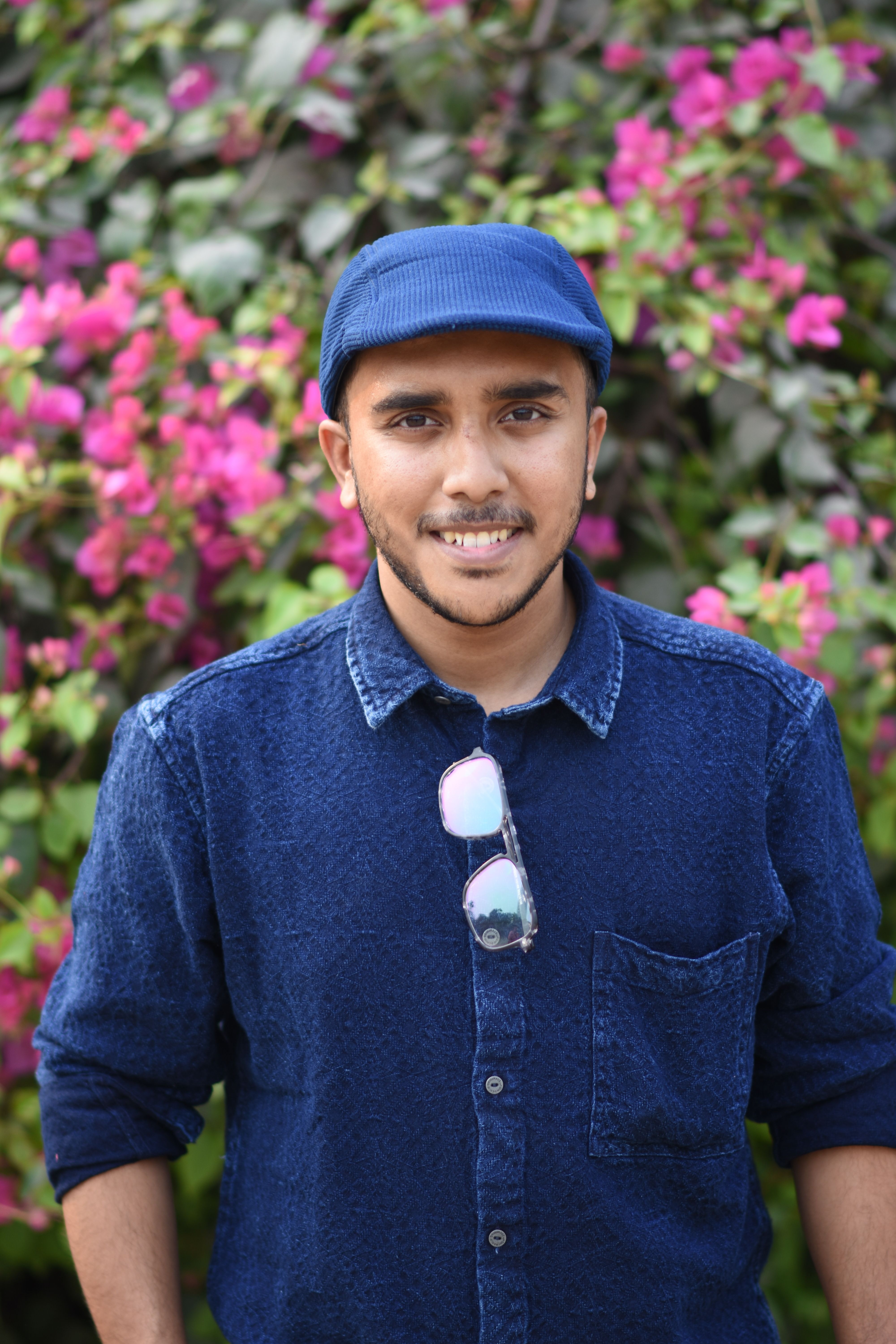 Smiling young man in a denim outfit with pink flowers behind, exuding casual charm.