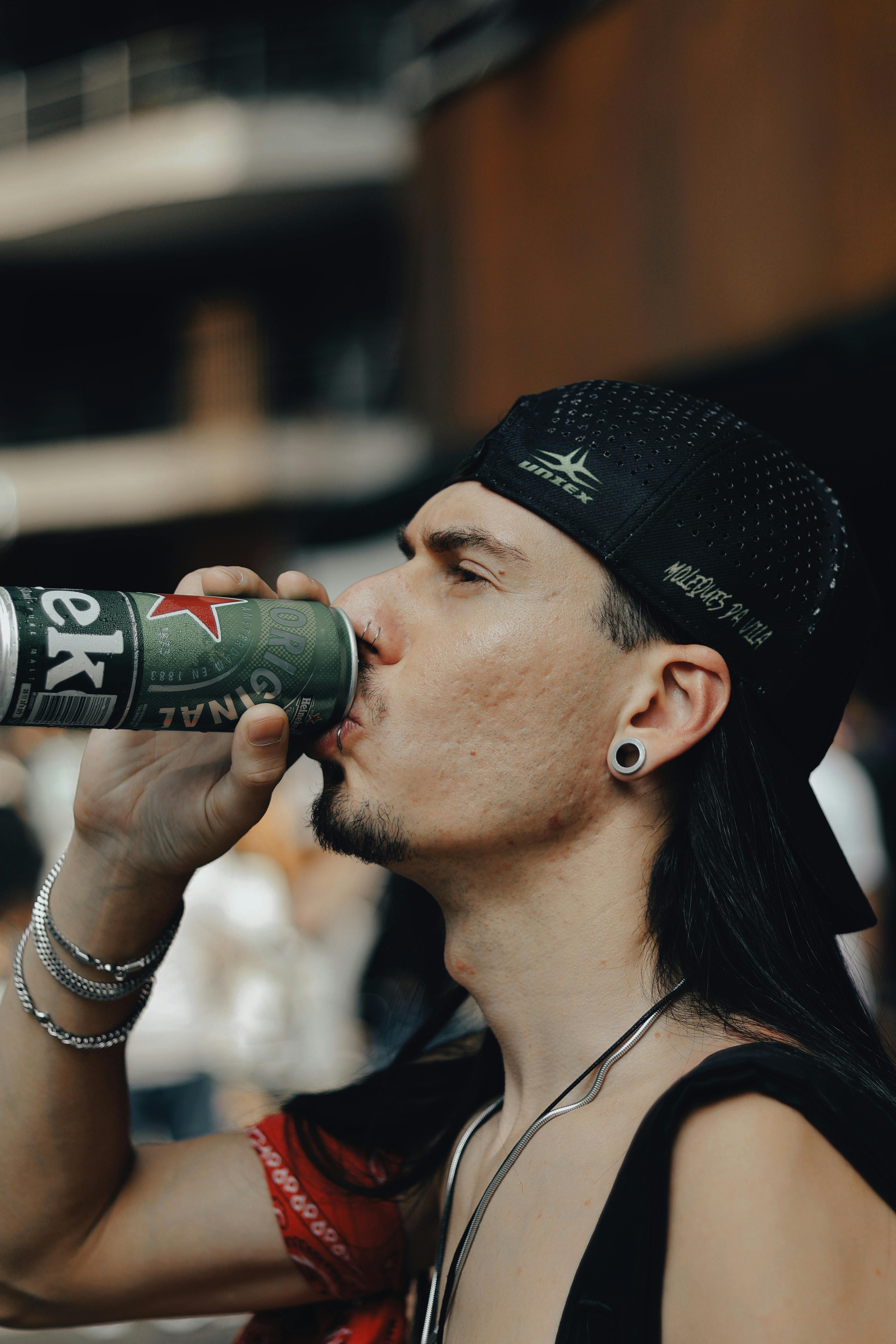Kostenlos Ein junger Mann mit Kappe und Ohrringen trinkt im Freien aus einer Dose und genießt seine Freizeit. Stock-Foto