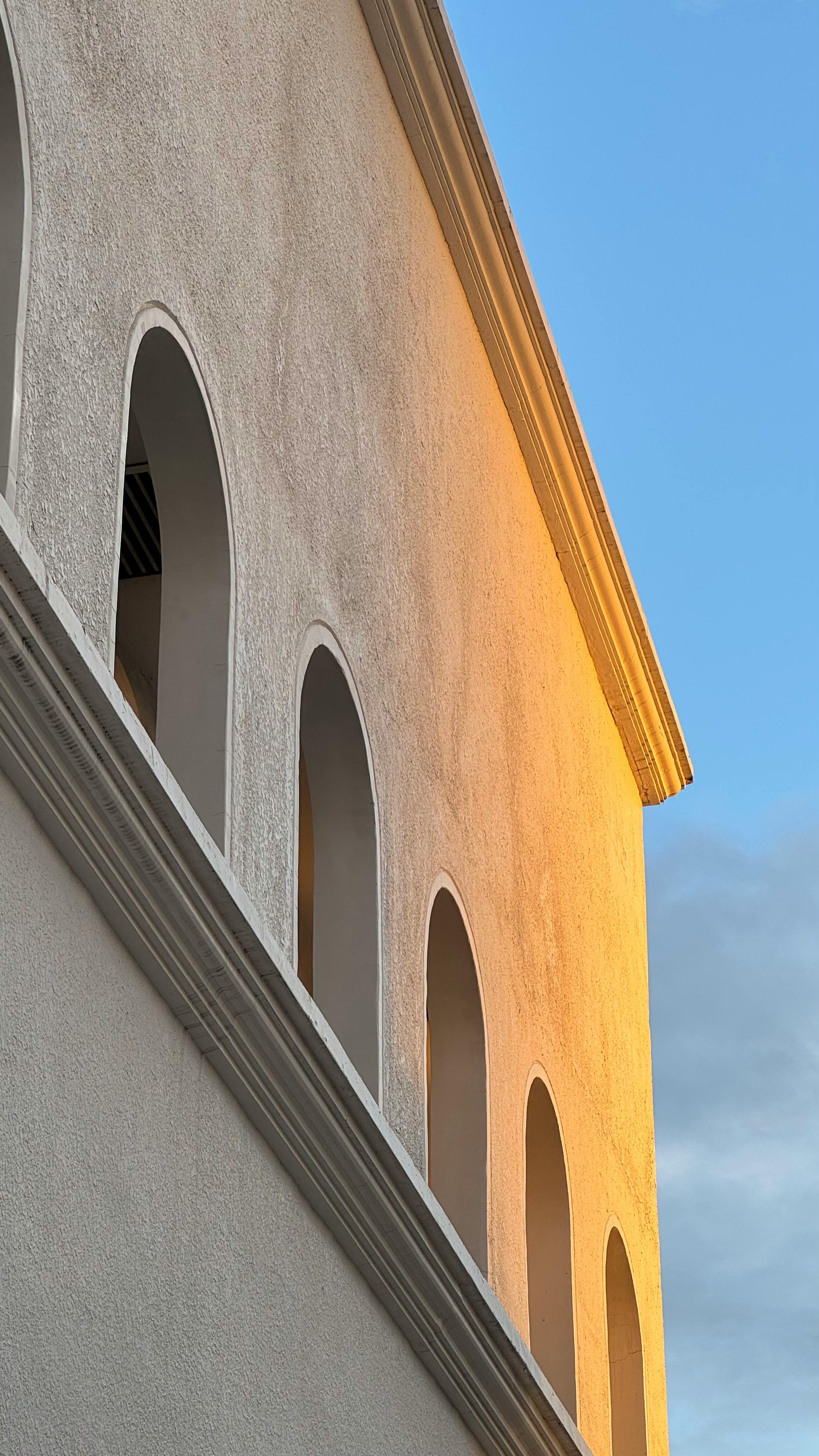 бесплатная Фасад здания, залитый солнечным светом, с арочными окнами, отражающими свет «золотого часа» на фоне голубого неба. Стоковое фото