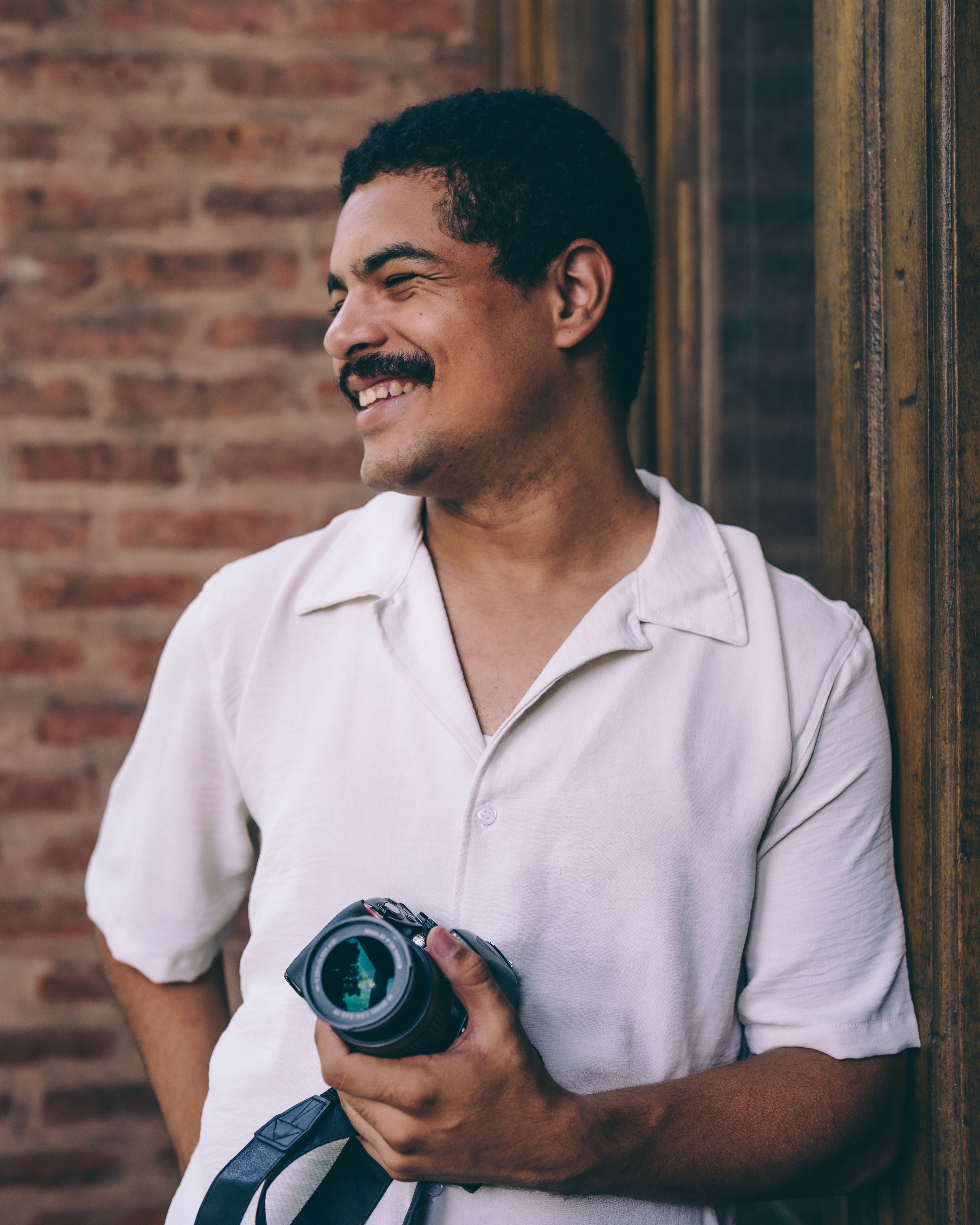 A joyful man holding a camera, standing beside a brick wall and window, exudes happiness.
