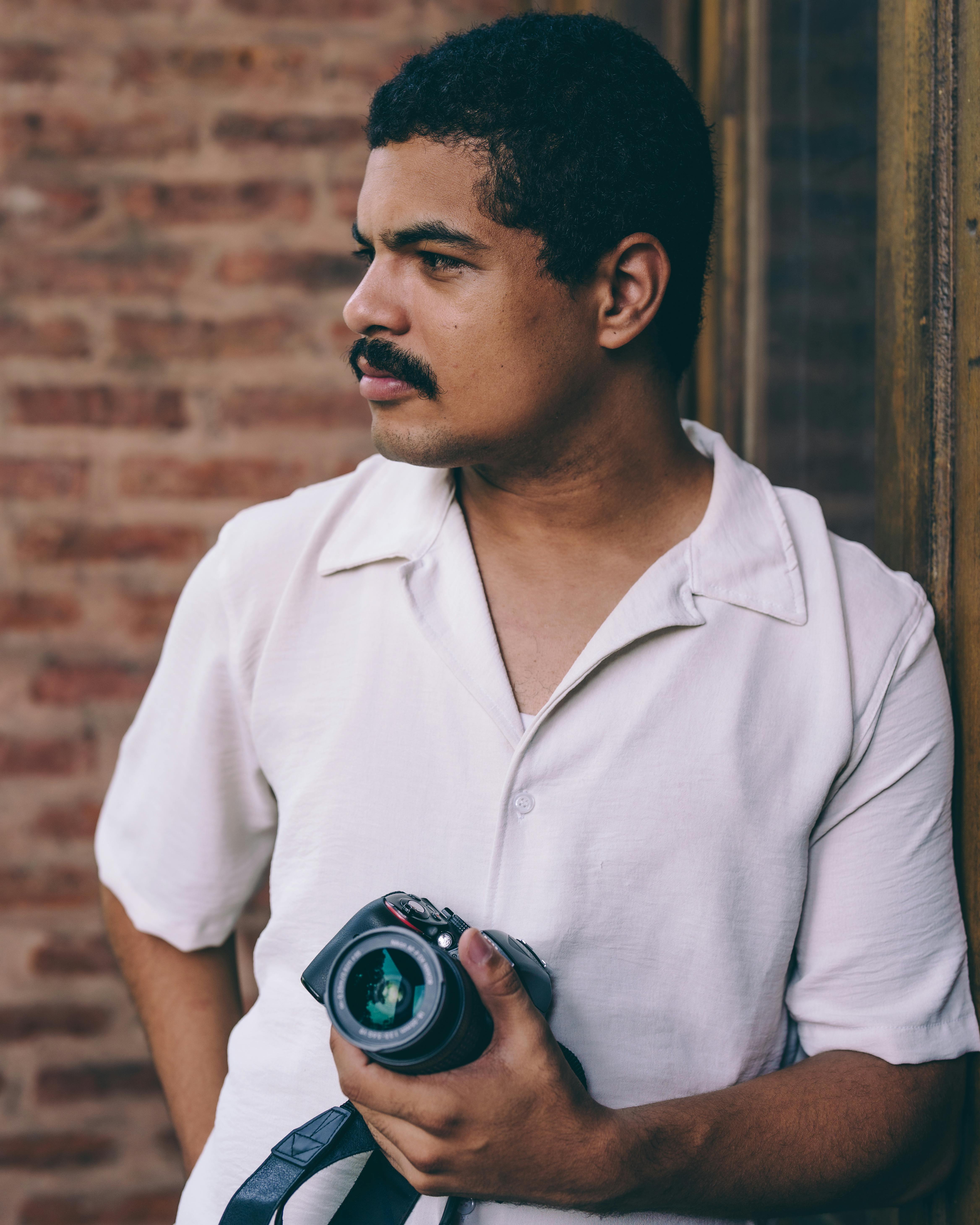 A thoughtful man holding a camera, leaning against a brick wall indoors.