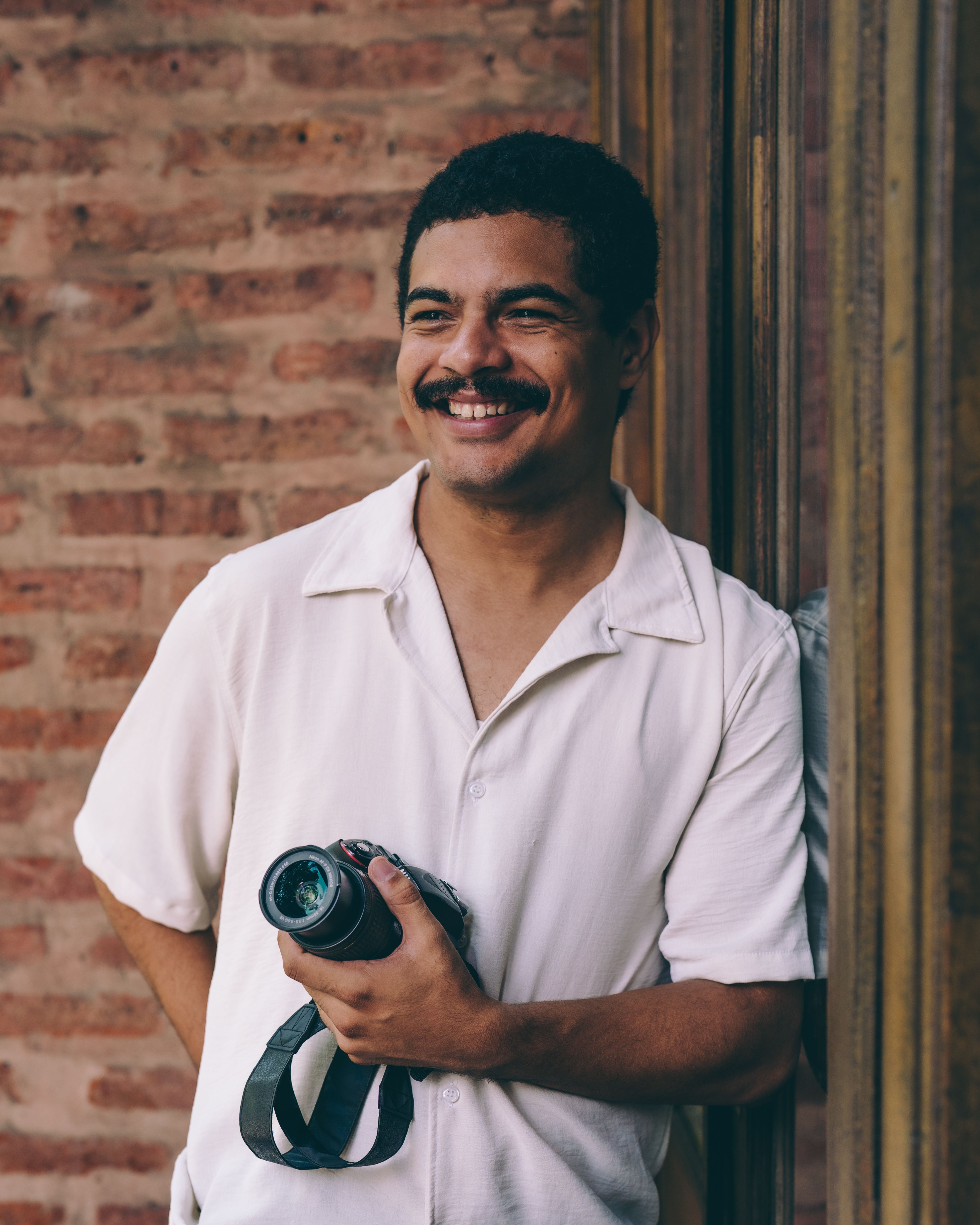 A cheerful photographer with a camera standing against a brick wall, enjoying the day.