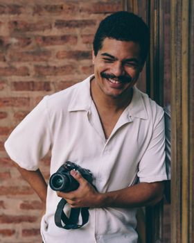 Portrait of a smiling photographer holding a camera against a brick wall.