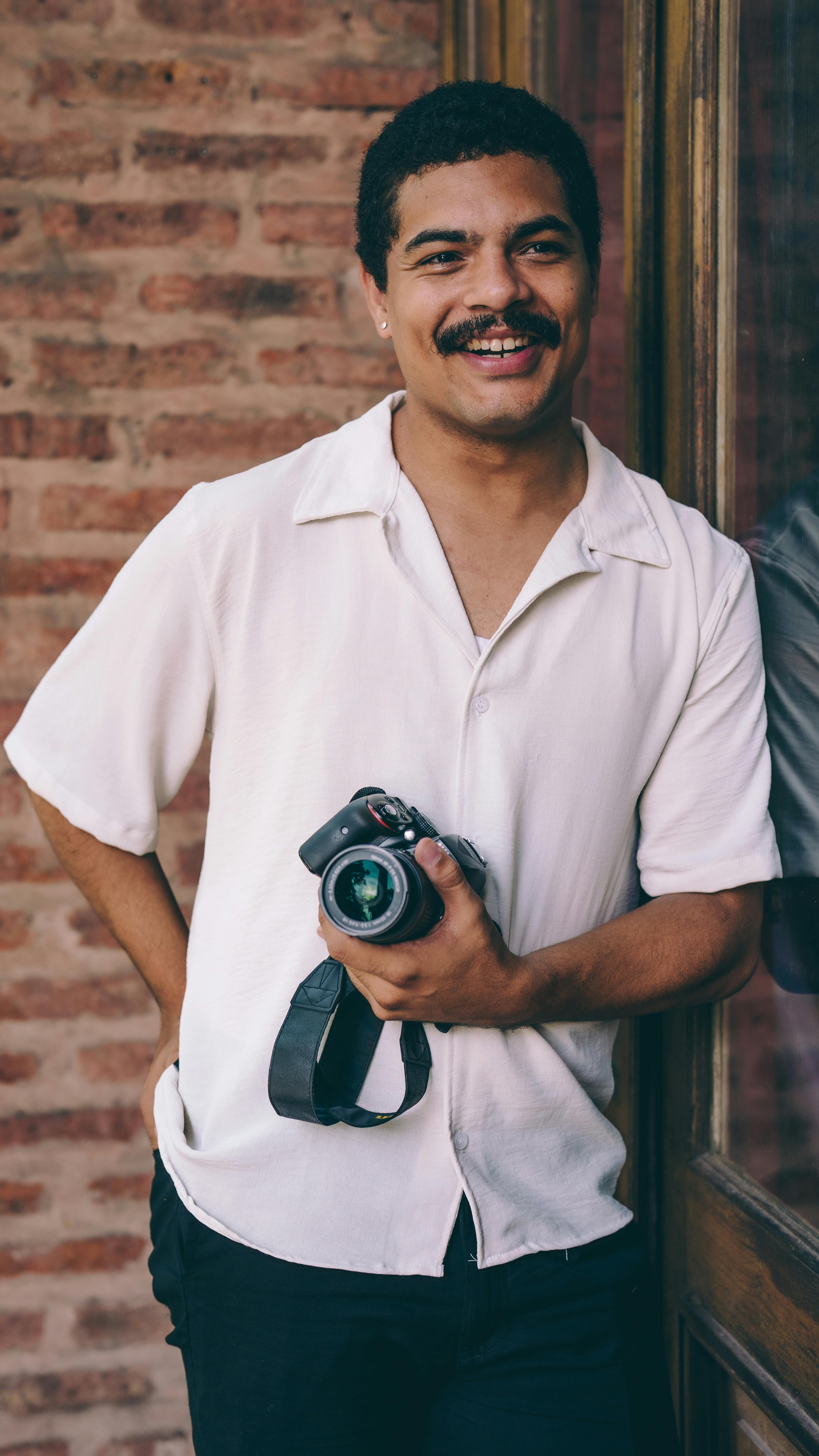 Portrait of a cheerful photographer holding a camera, leaning against a brick wall.