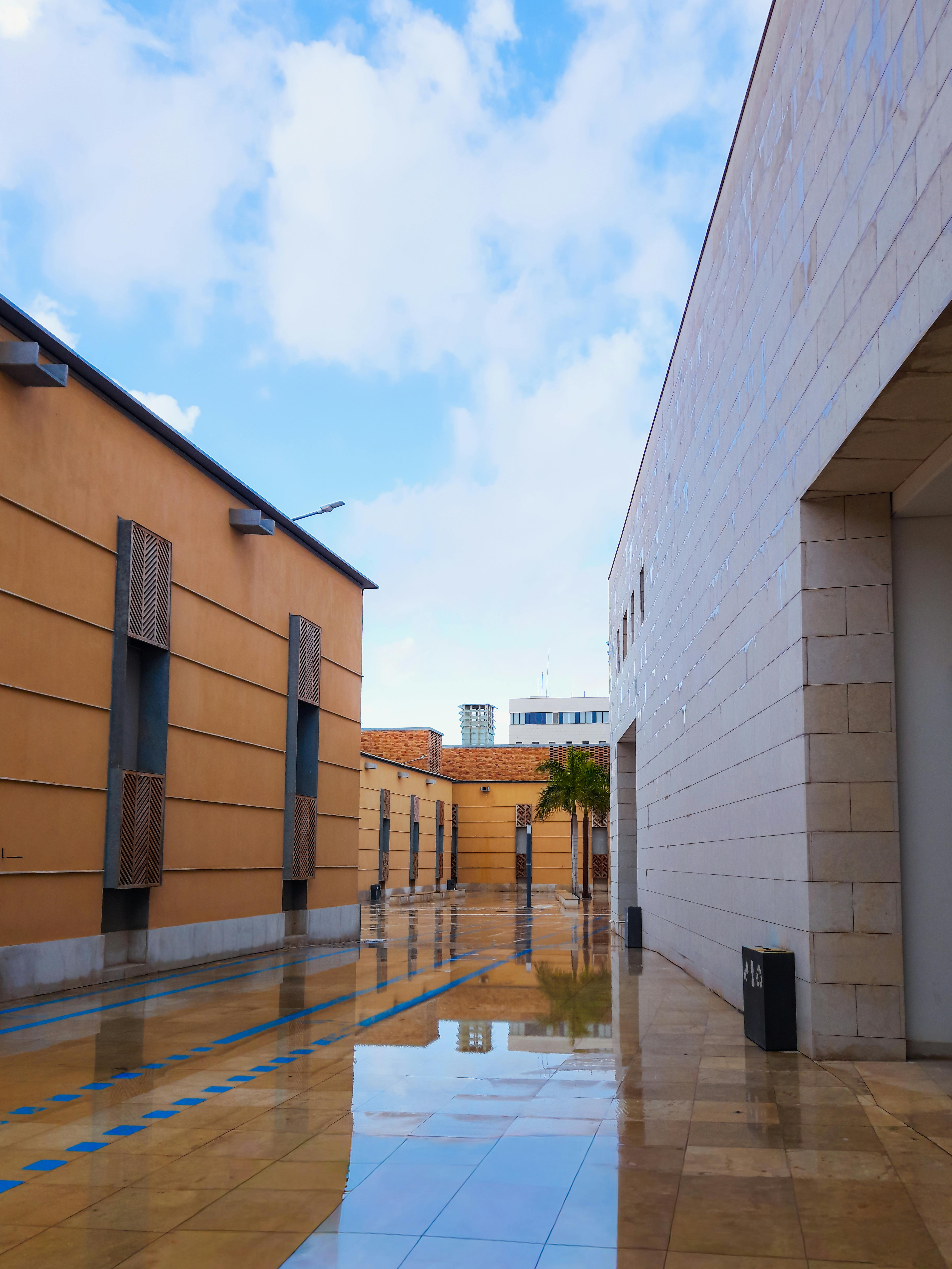 Gratis En moderne bygård med reflekterende våde fliser mellem moderne bygninger under en blå himmel. Lagerfoto