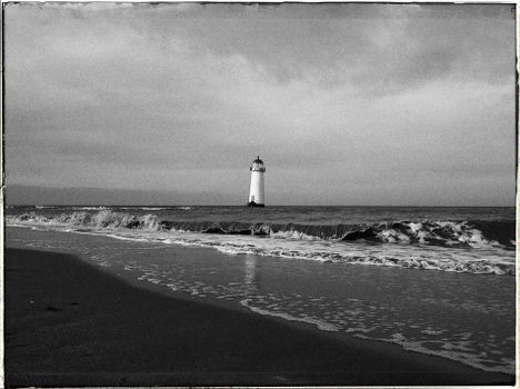 Black and white photo of a historic lighthouse standing on a serene beach with gentle waves.