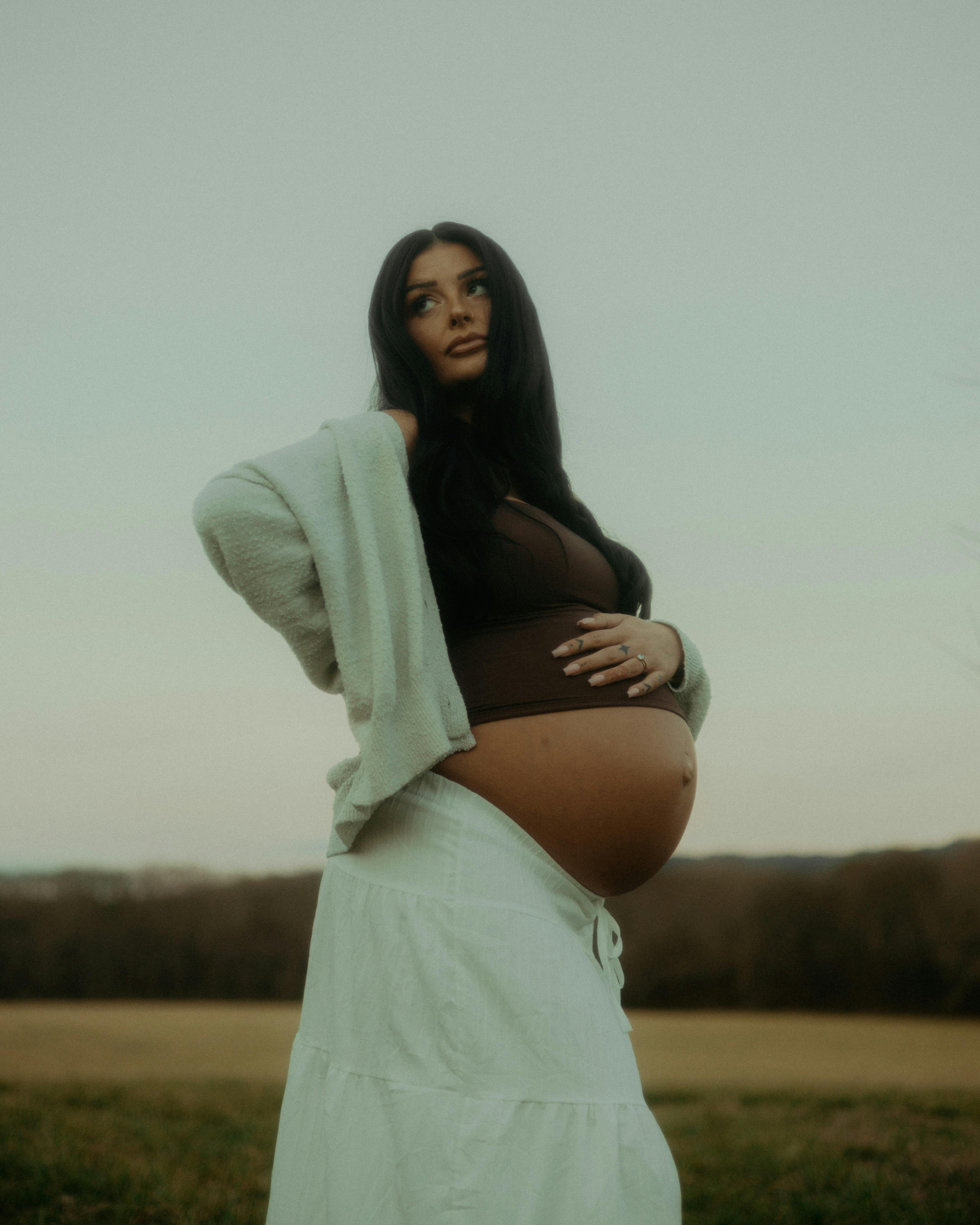 Kostenlos Eine schwangere Frau in einem Feld, bekleidet mit einem weißen Rock, einem braunen Oberteil und einer Strickjacke. Aufgenommen bei natürlichem Licht. Stock-Foto